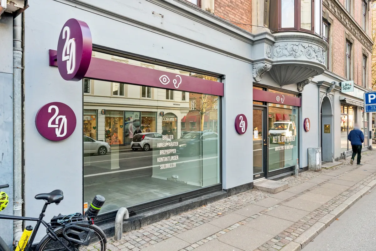 Menneske går forbi en butik med skiltet 2G, der tilbyder høreapparater, kontaktlinser og solbriller, og en cykel står foran vinduet