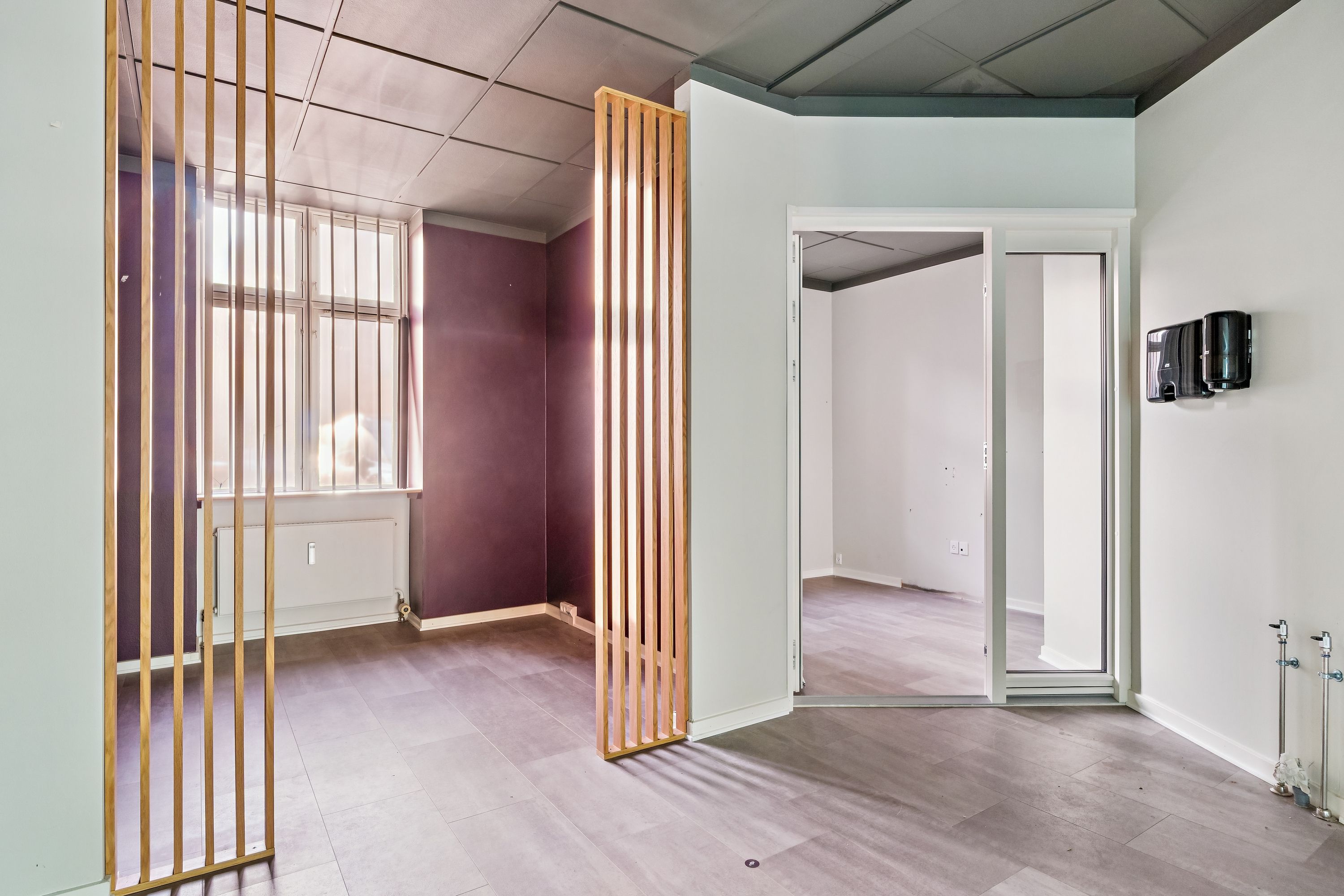 Indvendigt vinduesglas, gulv og hvide vægge, møbler mangler, åbne døre og toiletpapirautomater på væggen, højt loft og lys gulvbelægning