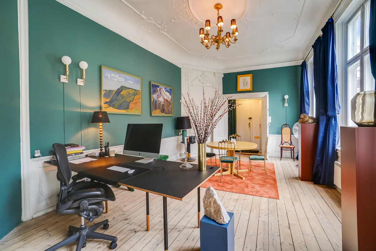 Kontor med computer, stol, skrivebord, lysekroner, gulvtæppe og stole i hyggelig baggård i København. Malerier og lysestager pryder væggene.