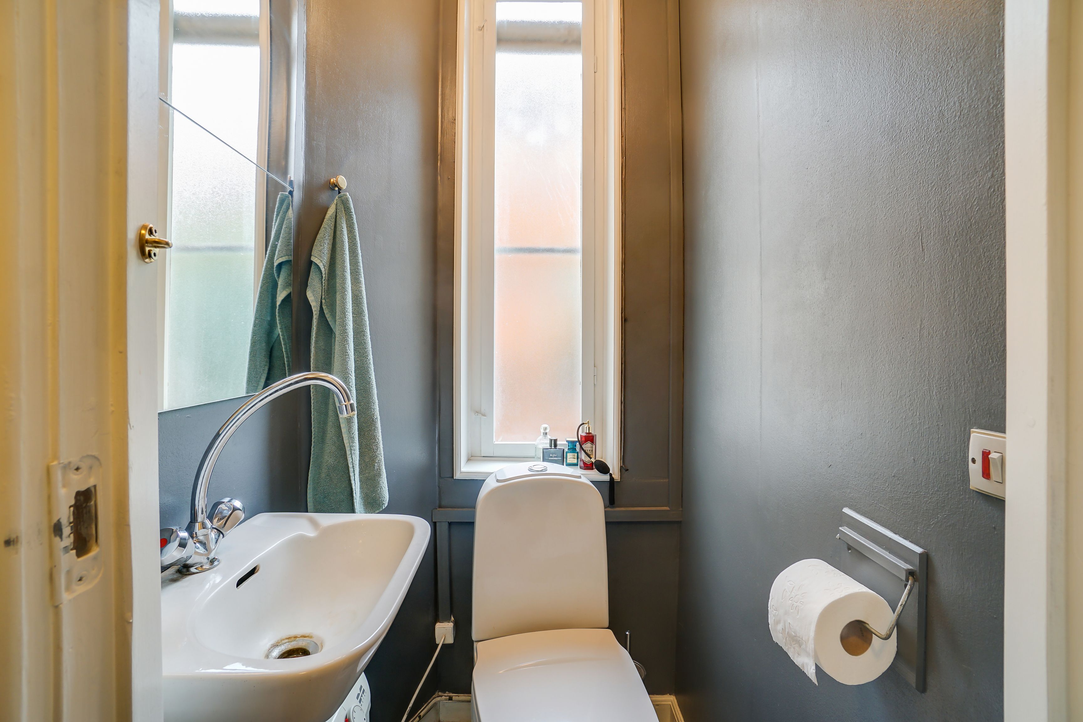 Lukket toilet med hvid toiletbøsse og hvidt vasketøj samt håndklæder og sæbe. Toiletpapirrulle i holder og en lille vinduesåbning med udsigt til baggården.