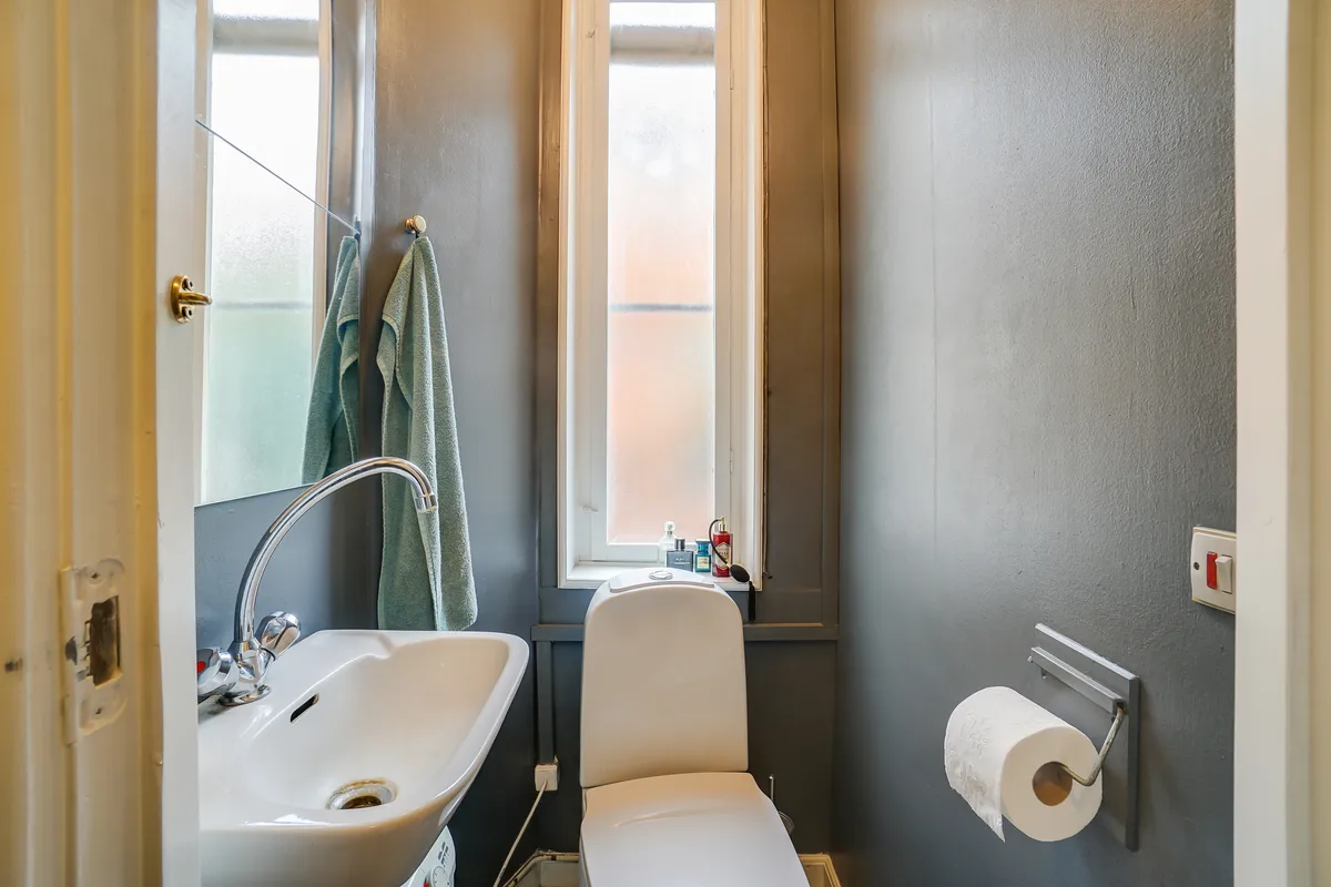 Lukket toilet med hvid toiletbøsse og hvidt vasketøj samt håndklæder og sæbe. Toiletpapirrulle i holder og en lille vinduesåbning med udsigt til baggården.