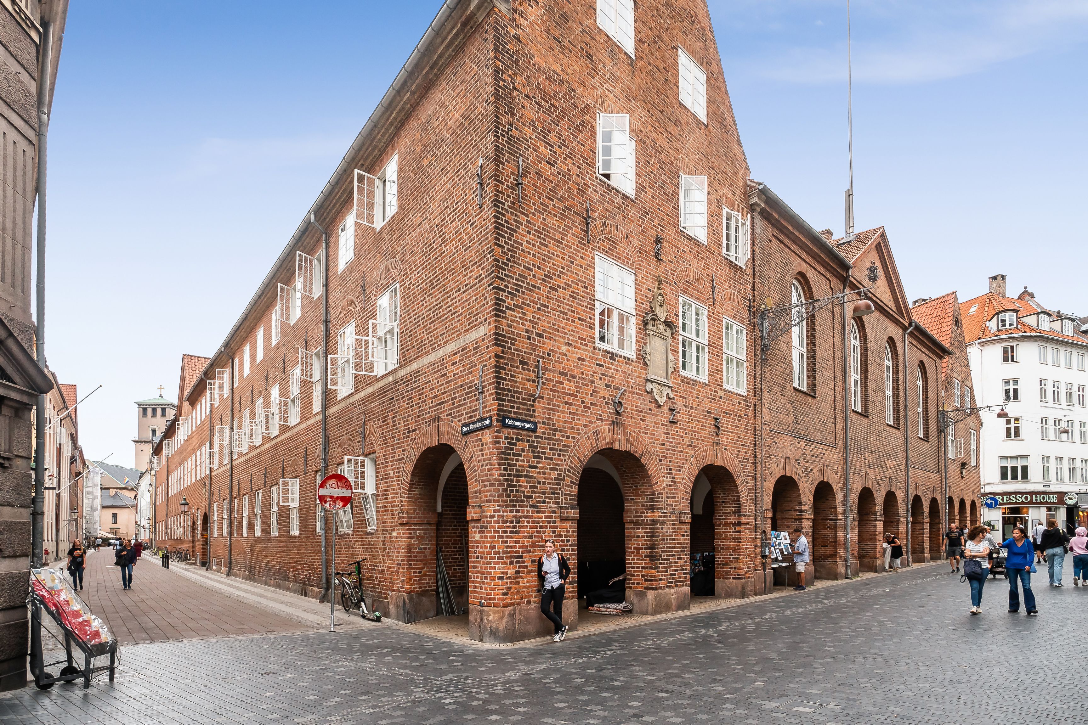 Et rødt murstenshus med flere vinduer og buer, udenfor går folk og står folk og en cykel står parkeret