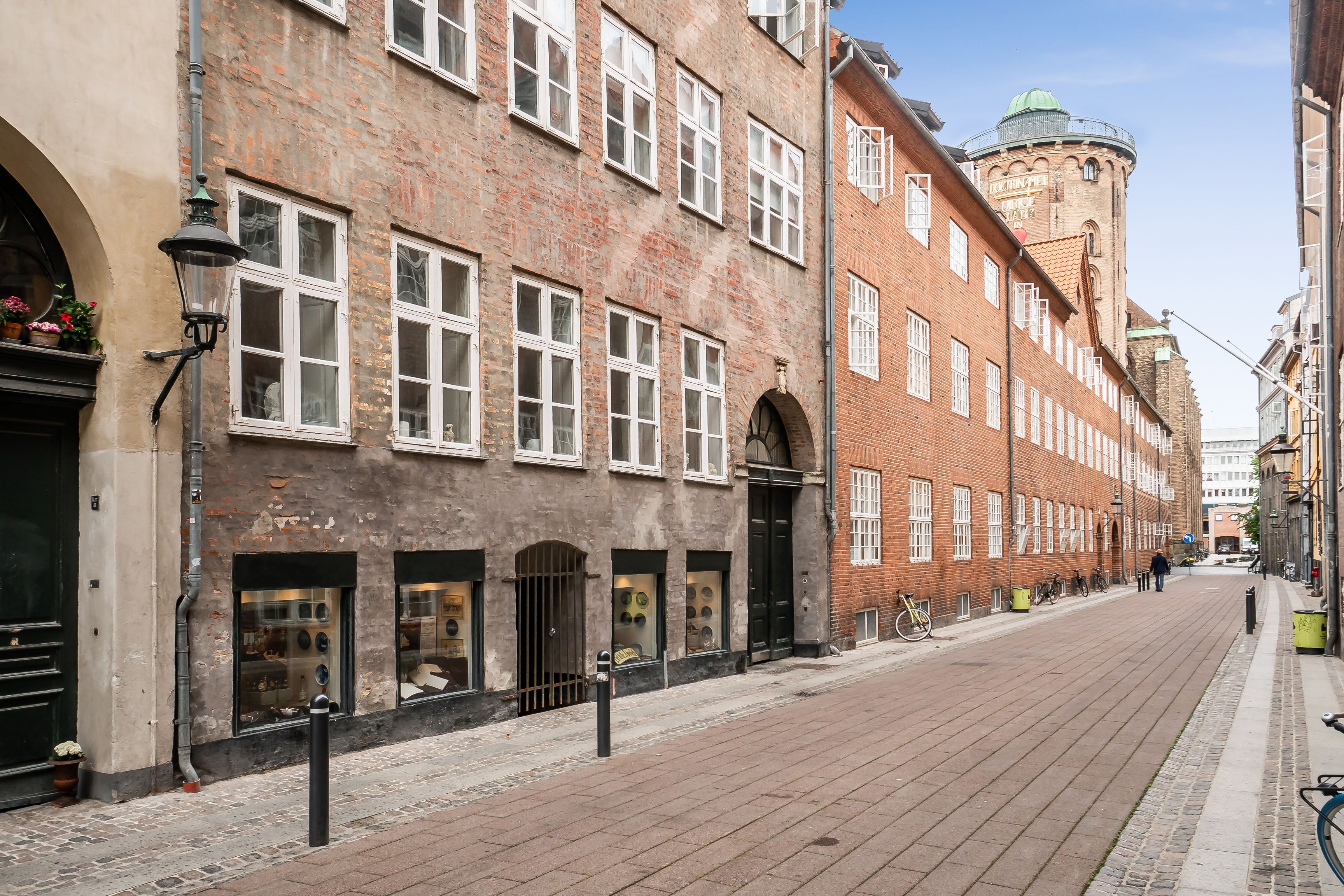 En fredelig gade med gamle bygninger, røde mursten og store vinduer. Butikker med glasruder og nogle blomster. En person går langs en tom gade med gamle gulvsten og søjler.