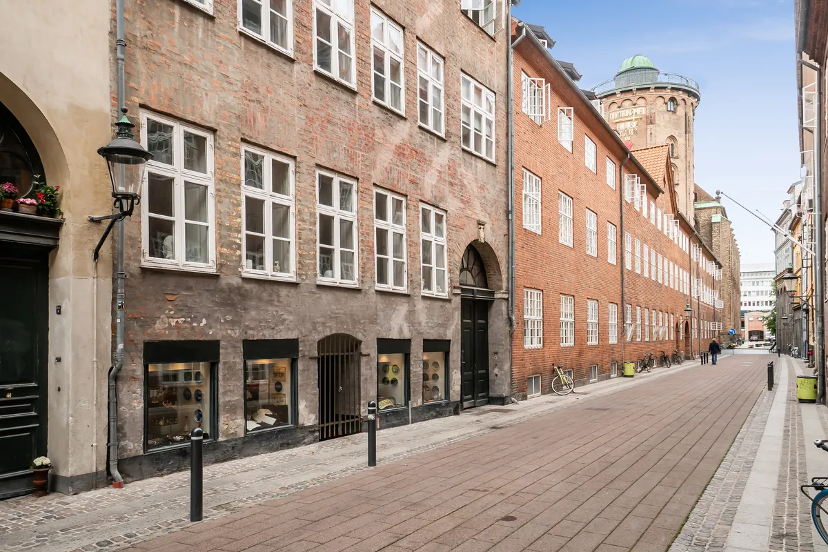En fredelig gade med gamle bygninger, røde mursten og store vinduer. Butikker med glasruder og nogle blomster. En person går langs en tom gade med gamle gulvsten og søjler.