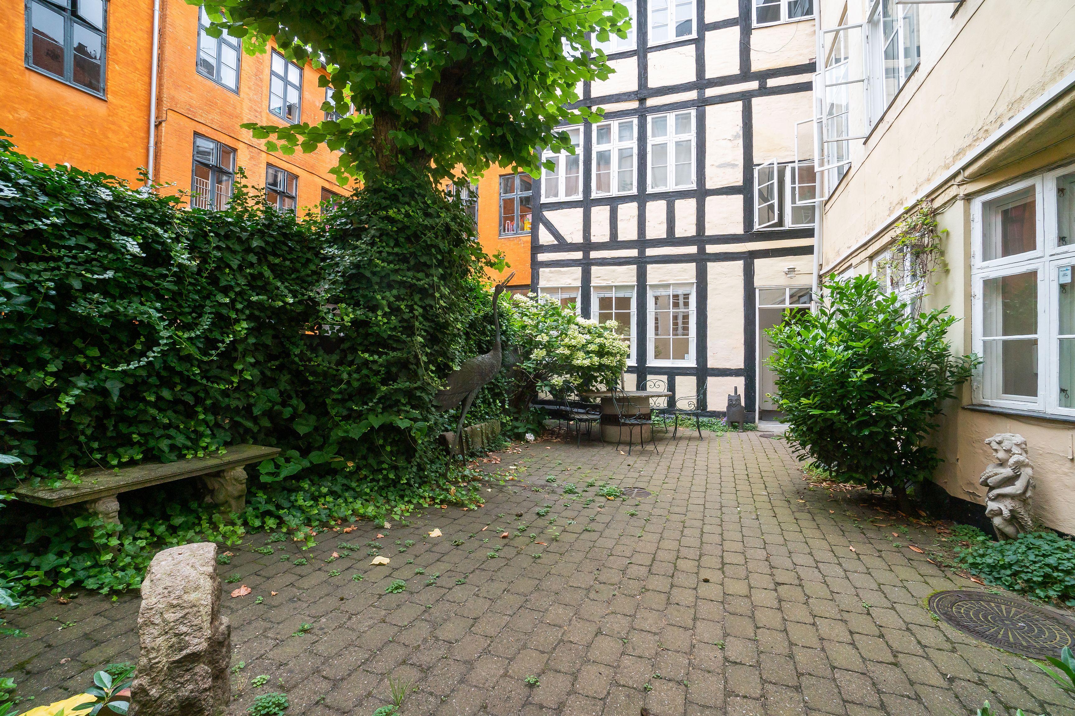 Baggård med terrasse, træer, buske og statuer, stenbenk og skur, røde og hvide bygninger med vinduer, lyse og skyggede områder, terrassemøbler og udsigt til gade.