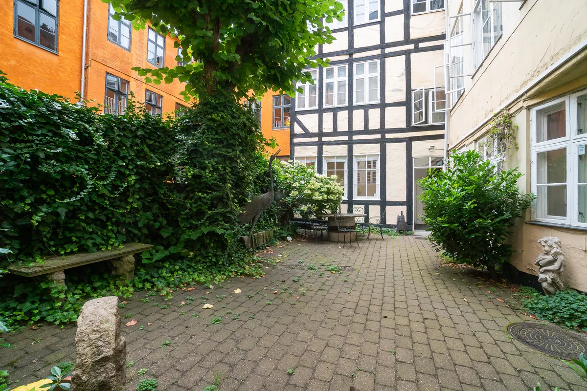 Baggård med terrasse, træer, buske og statuer, stenbenk og skur, røde og hvide bygninger med vinduer, lyse og skyggede områder, terrassemøbler og udsigt til gade.