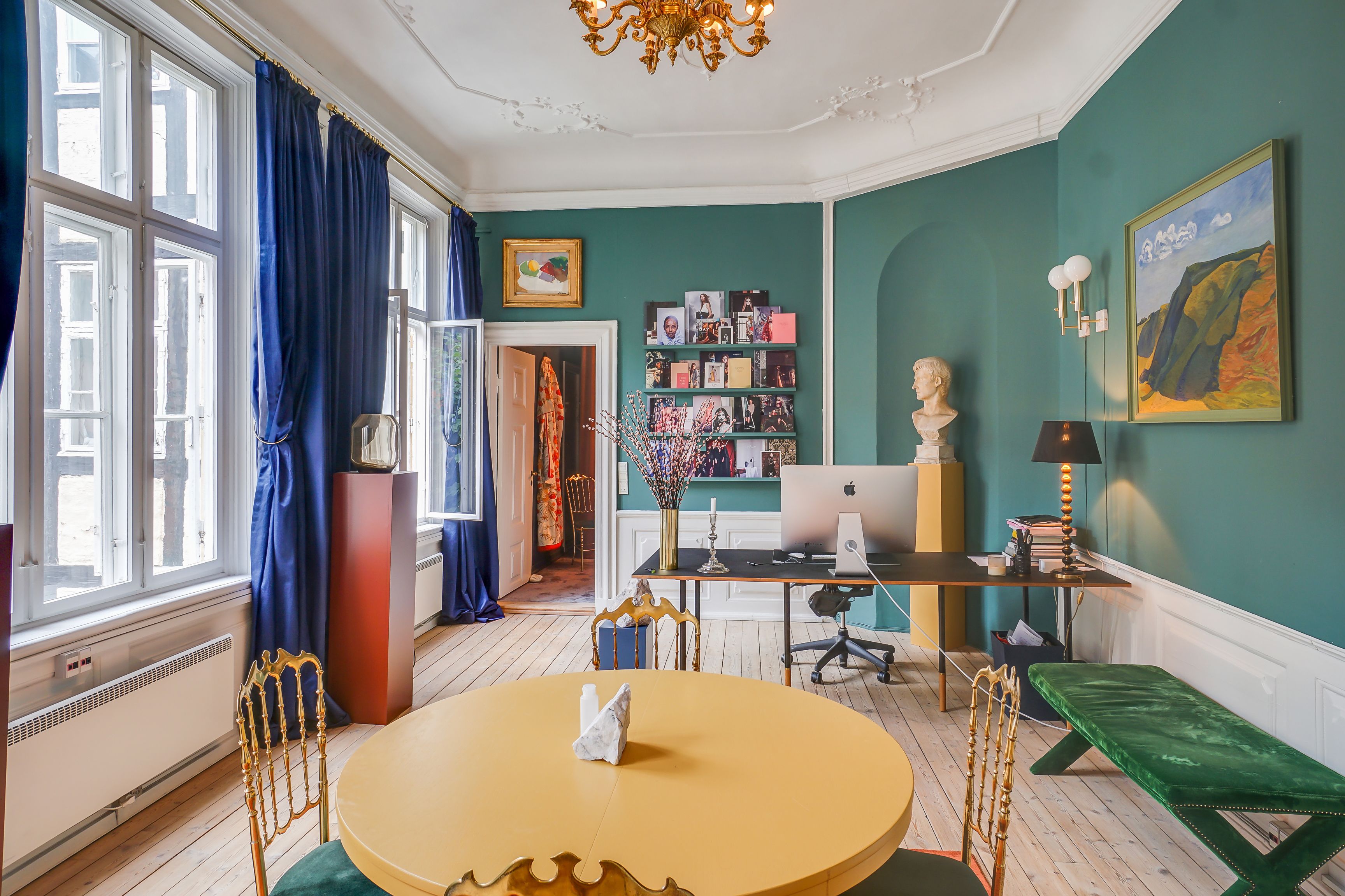 Kontor med gult rundt bord og guldfarvede stole, grønne vægge, lysekroner, malerier, skrivebord, computer, bue og lysestage samt åbne vinduer med blå gardiner