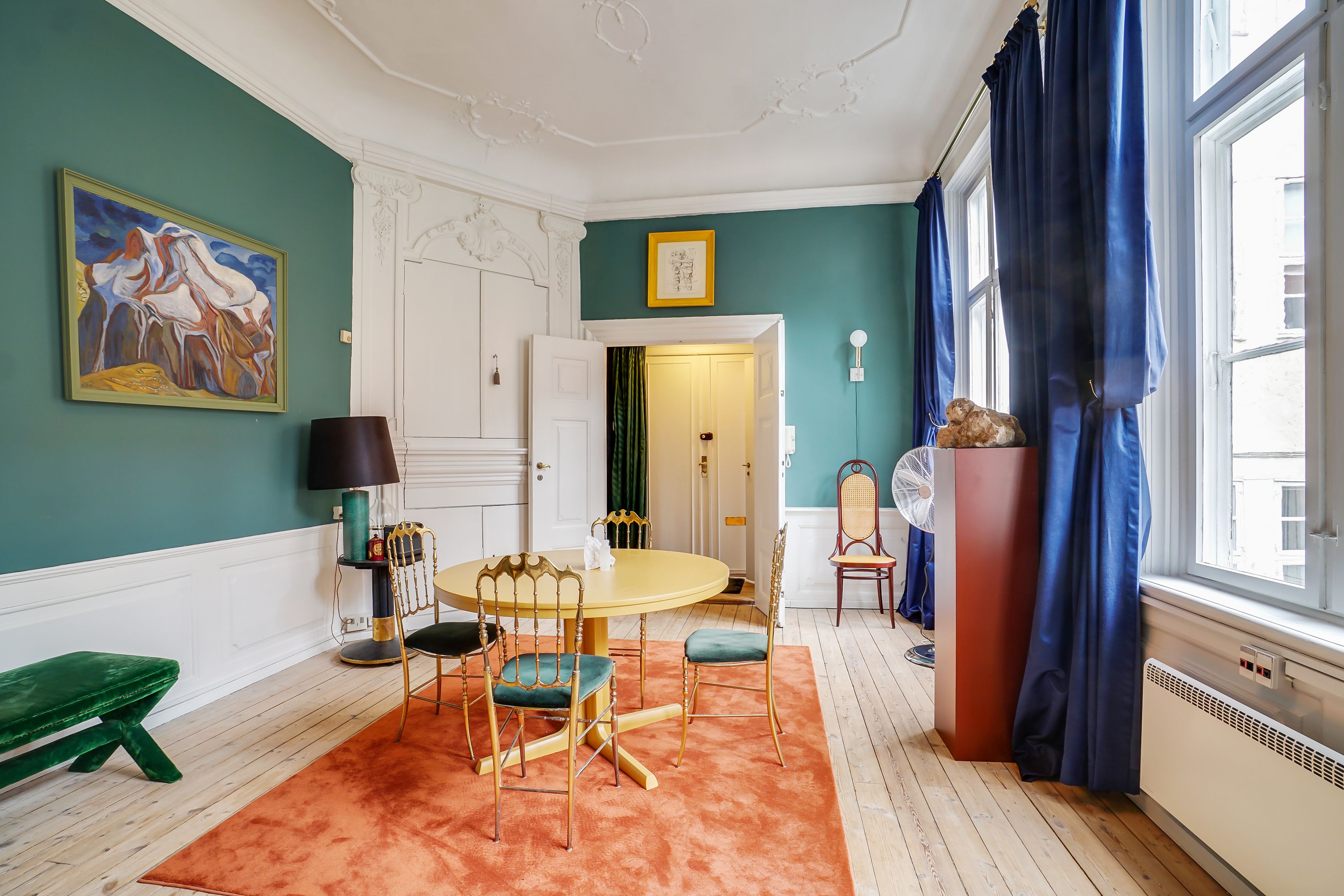 Kontor til leje med smuk gulvtæppe, lysegrå vægmalerier, gult rundt bord med guldstole og lysegrå vægmalerier med en malet bjerglandskap og en lysegrå væglampe.