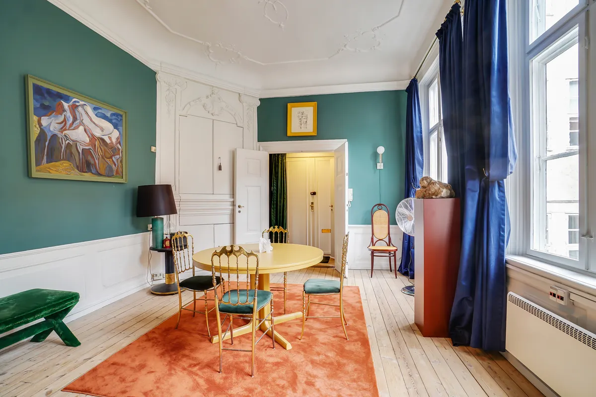 Kontor til leje med smuk gulvtæppe, lysegrå vægmalerier, gult rundt bord med guldstole og lysegrå vægmalerier med en malet bjerglandskap og en lysegrå væglampe.