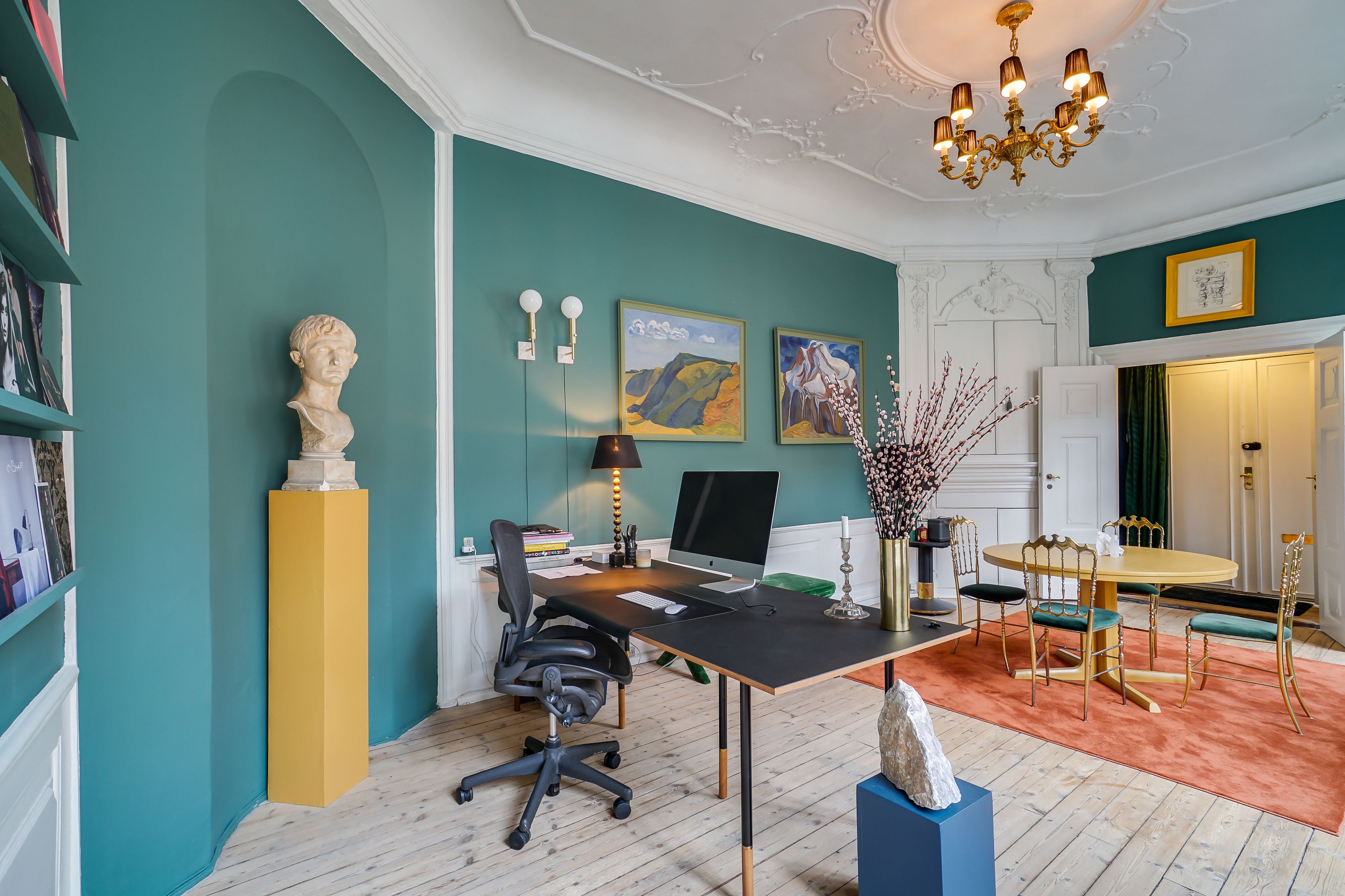 Kontor med gulvvarme, skrivebord, stol, buste, skrivebordslampe, computer og gulvtæppe i lysegrå stue med tegninger og lysekroner