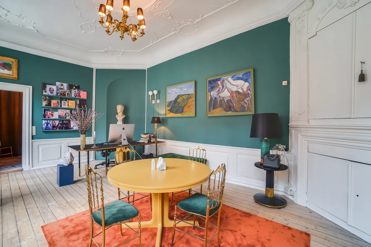 Kontor med grønne vægge, gul rund bord, og guldmøblerede stole. Væg med malerier, skrivebord med computer og vase. Gulvtæppe, lysekroner og lysekroner.