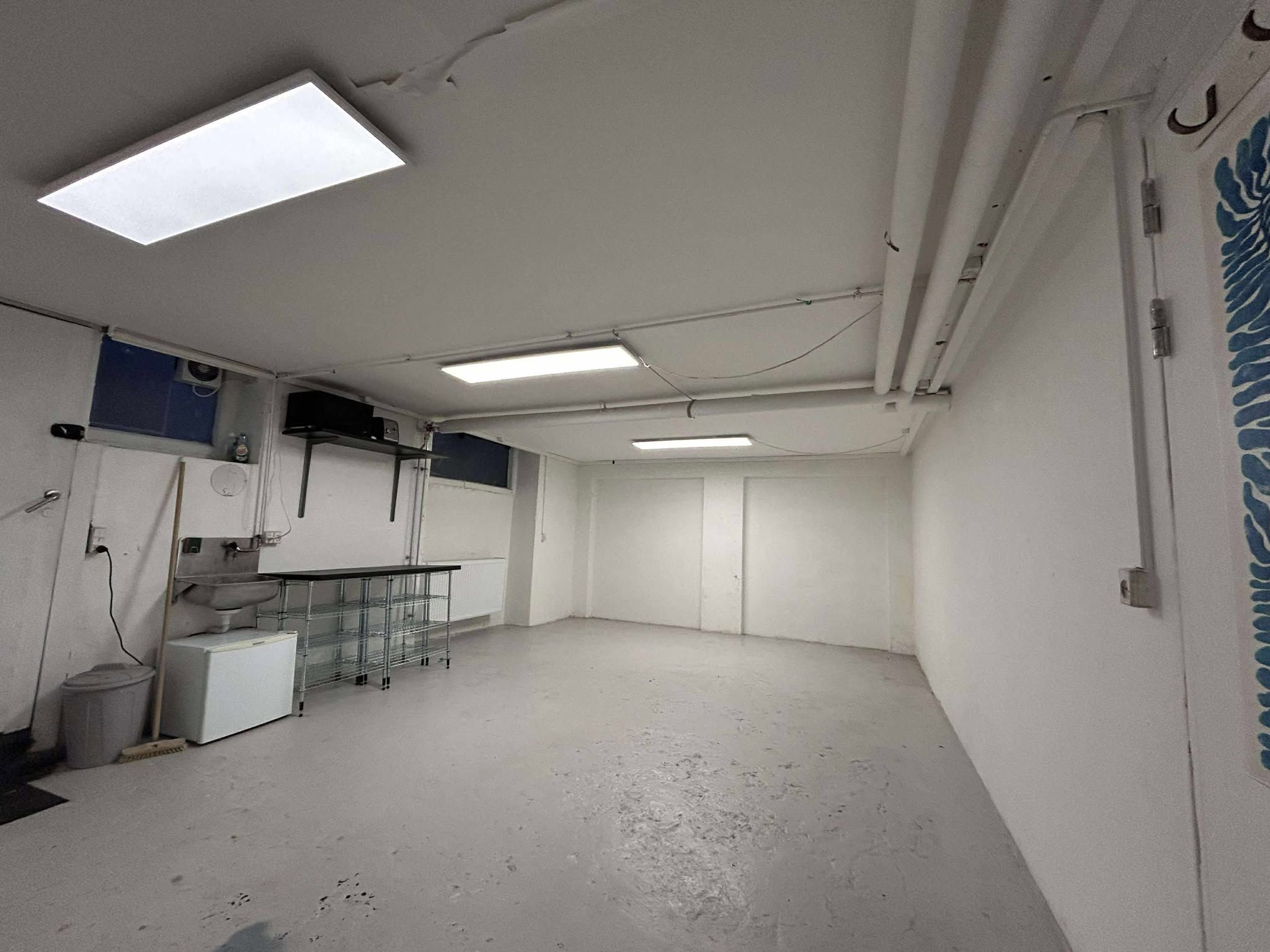 Kælderlokale med egen indgang og vinduer til leje i Frederiksberg. Lokale med radiator, køleskab og bord. Forskellige lysekroner er installeret på loftet.