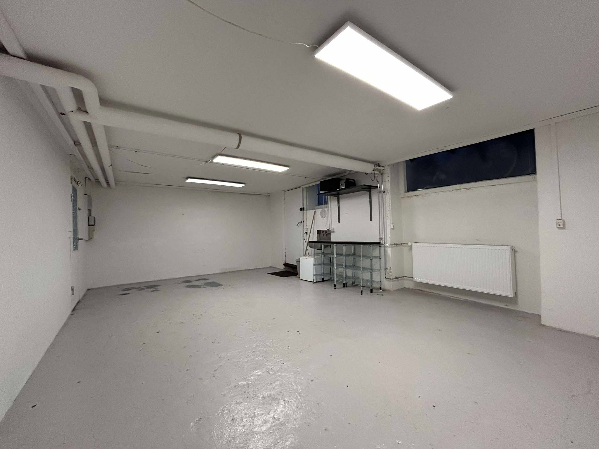 Kælderlokale med hvide vægge og gulv samt tre lysekroner. Et lille skrivebord med et hvidt køleskab og en stående radiator. Et vindue og et lukket skab med et tv.