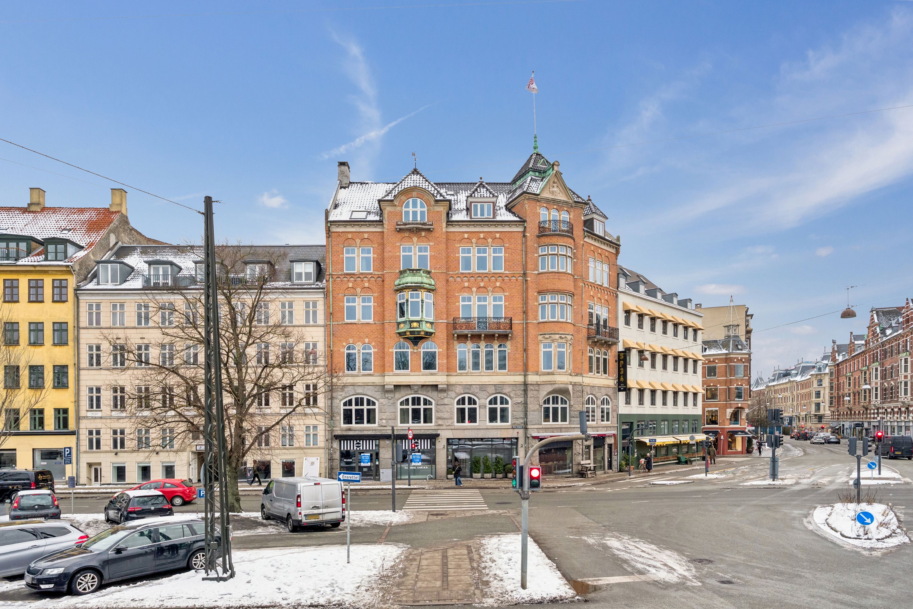 En butik på hjørnet af en gågade i København K med røde bygninger og biler parkeret langs siderne af vejenen og et stoplys i grønt