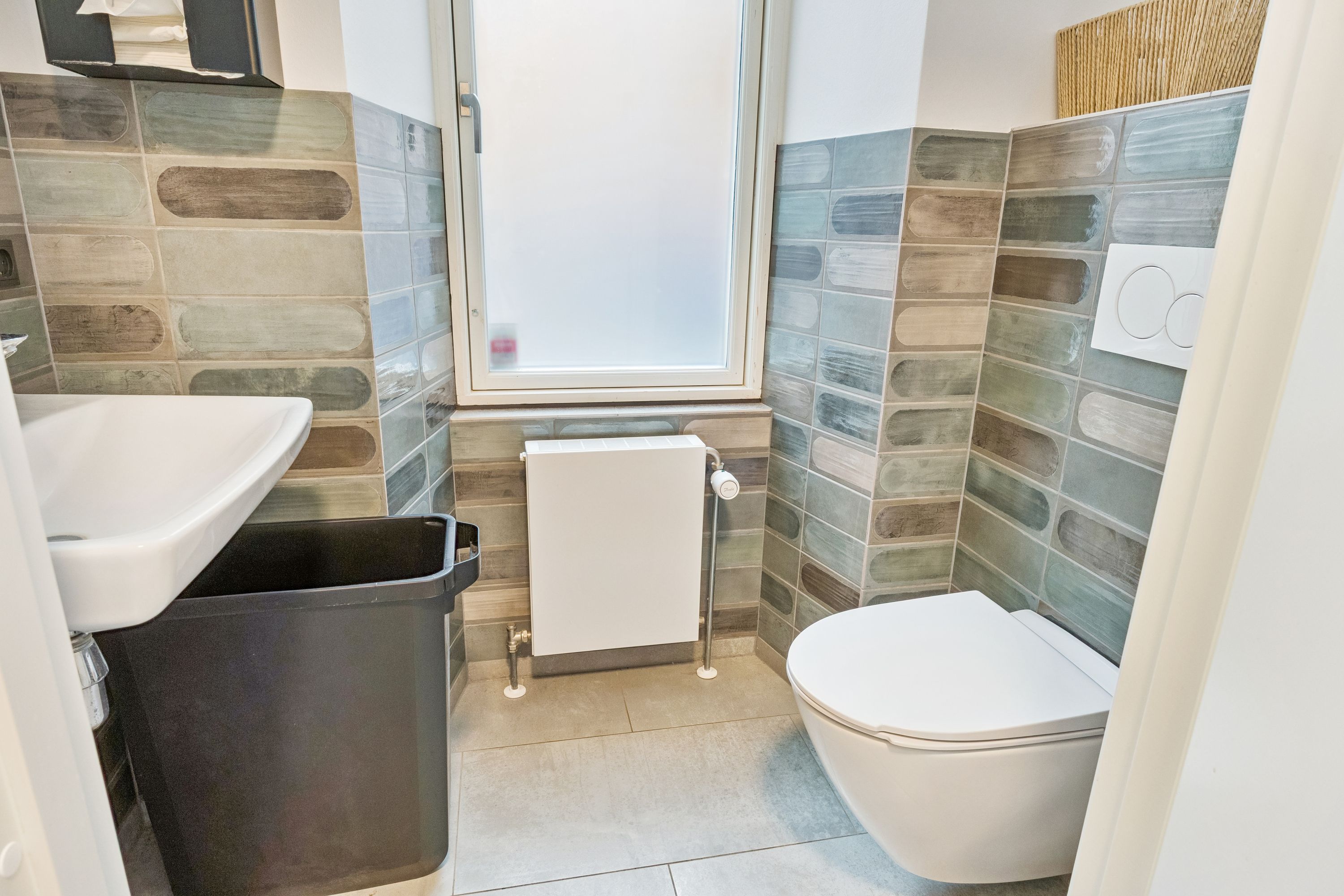 Et lille badeværelse med hvidt toilet, hvidt vasketøjsskab og hvidt badekar. Der er en ventil til vandvarmeren, og badeværelset er dekoreret med grønne og brune klinkefliser.