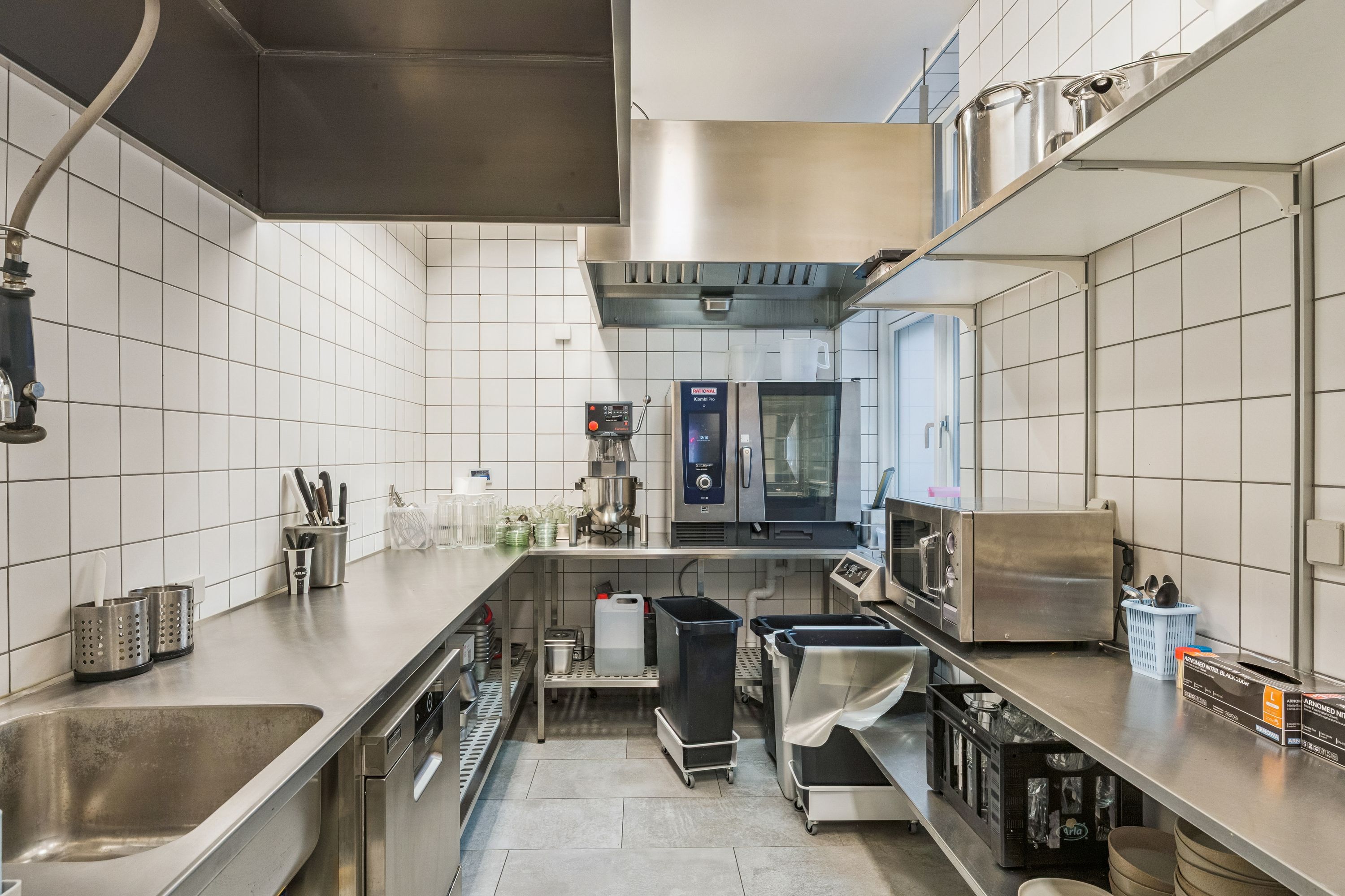 Køkken med moderne indretning og højkvalitetsapparater. Væggene er klædt med hvide flise og der er en stor metalborte med apparater. Et sæsonelt køleskab og en mikroovn står på højre side.