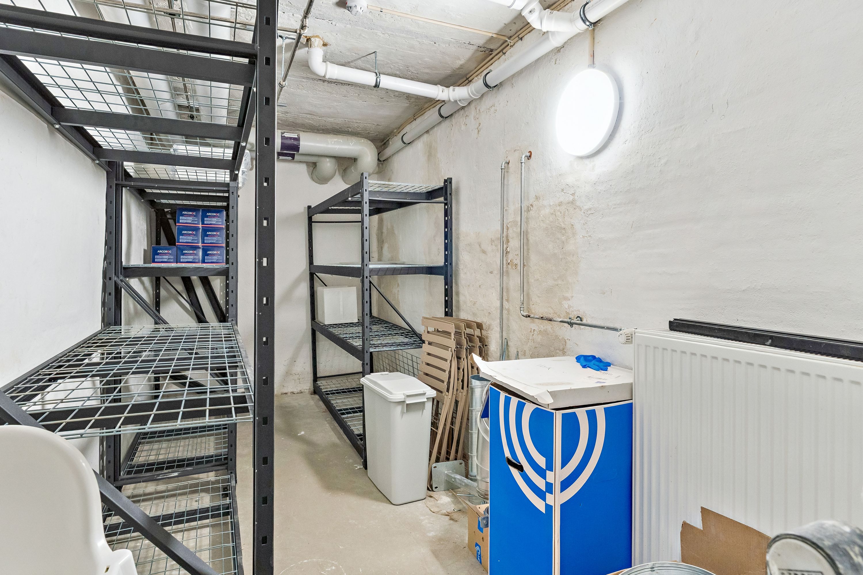 Lager med stålregaller, skraldespand, papkasser, hvid radiator og lyse pibesystemer. Værelse med højt til loftet og lysepunkter på væggen.