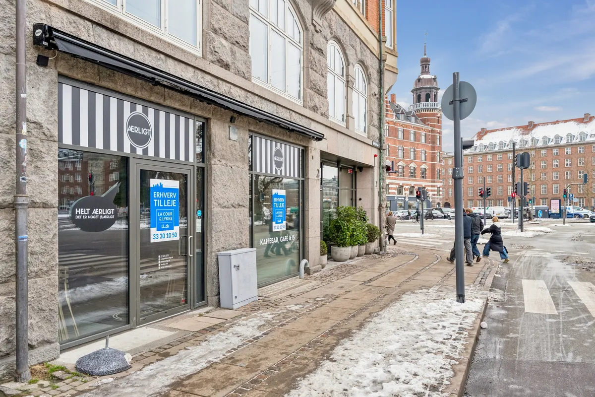 Cafe til leje på Nørre Voldgade, st. tv. I baggrunden er der en kirke, og adskillige biler er parkeret i området. Nogle mennesker går på fortovet.