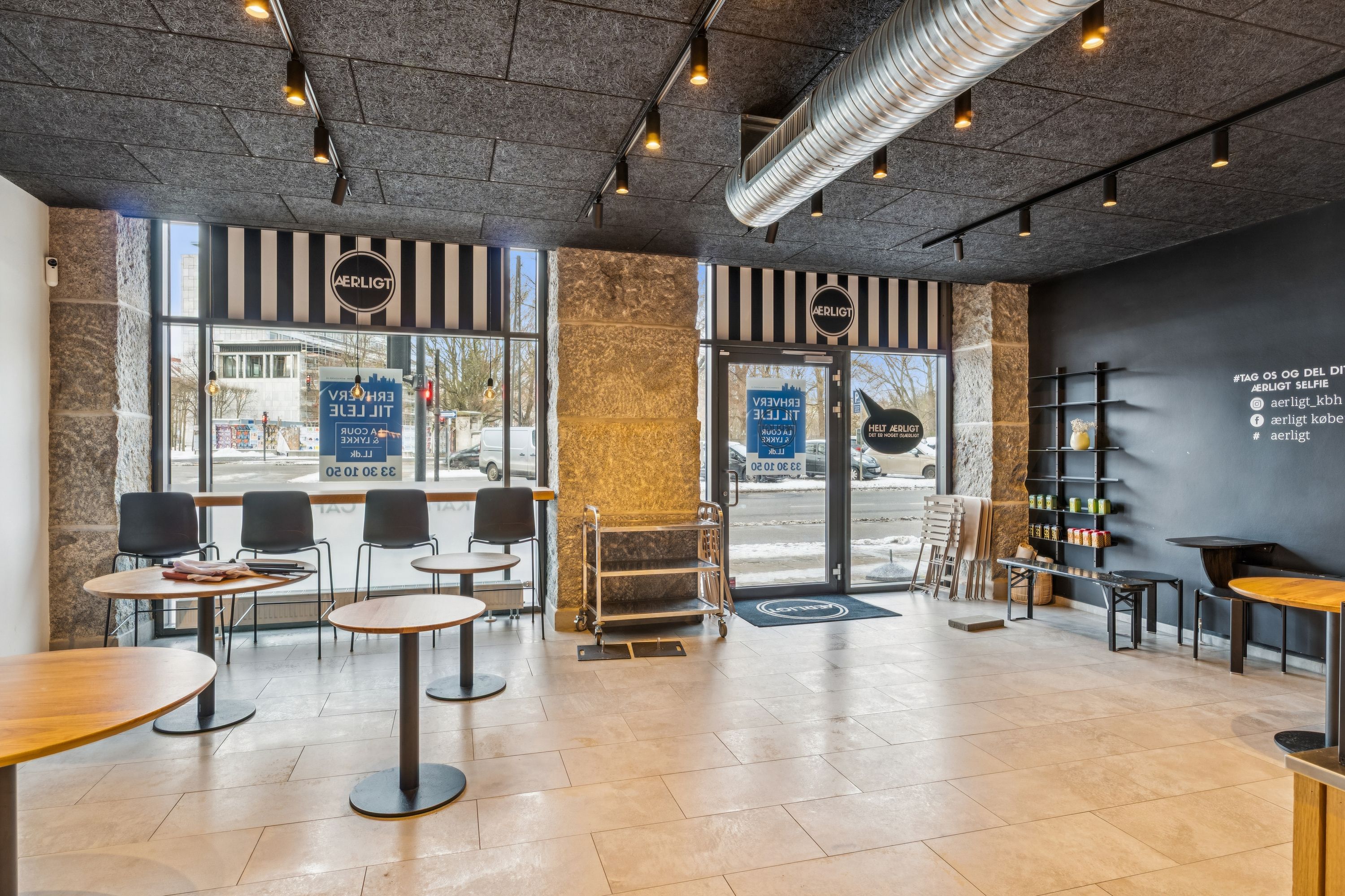 Ny café til leje i København K. Indvendigt område med runde borde og stole samt hylder med forskellige varer. Dør med skilt og vinduer med reklamer
