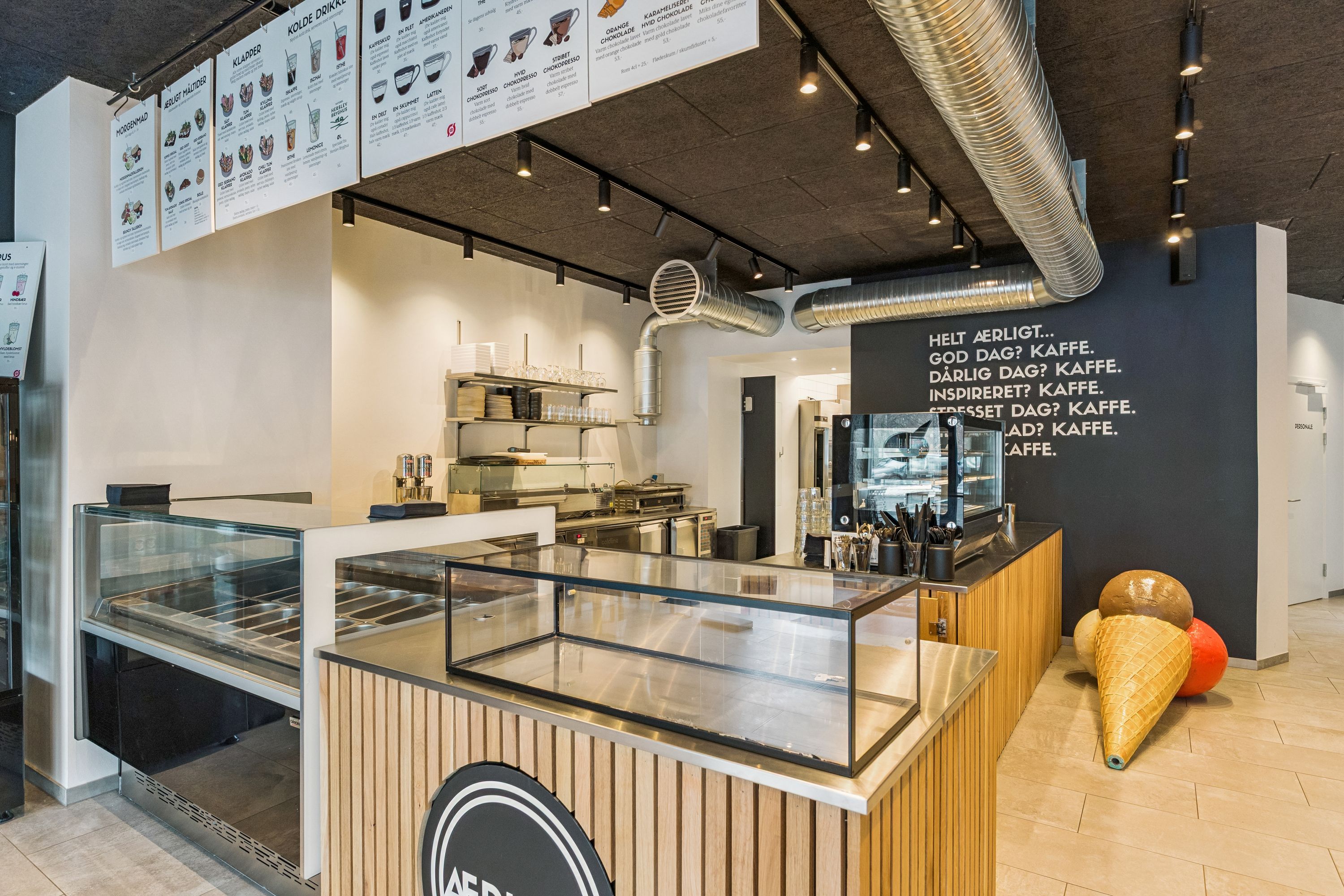 En ny café med åben køkken, serveringsdisk og udstillingsskabe. Der er tekst på væggen og iskæmme med smagfulde is. Der er højttalere og røgsuger på loftet