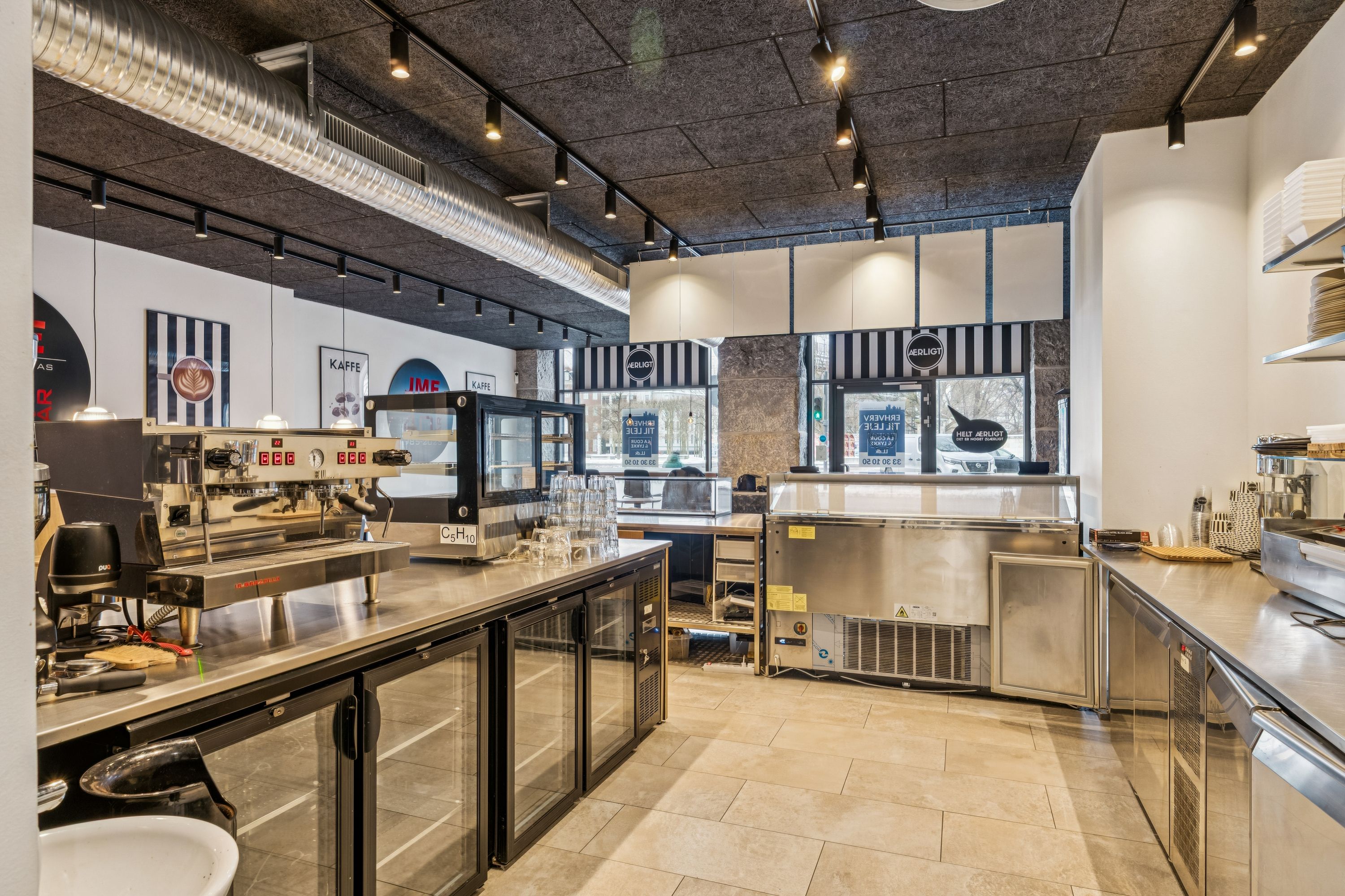 Inde i et moderne køkken er der en maskine, der ser ud som en espressomaskine. Væggene er hvide, og der er flere apparater og en stor ovn. Loftet er højt med flere lyspærer