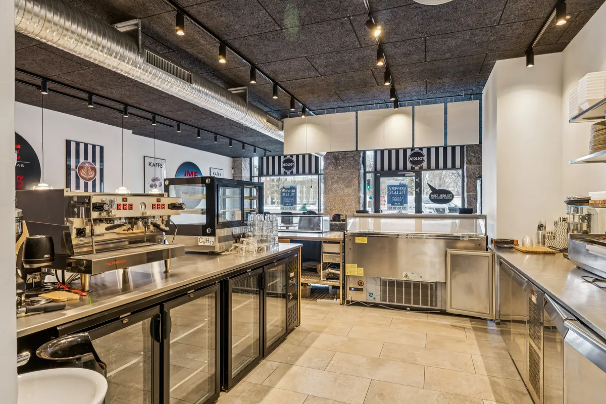 Inde i et moderne køkken er der en maskine, der ser ud som en espressomaskine. Væggene er hvide, og der er flere apparater og en stor ovn. Loftet er højt med flere lyspærer