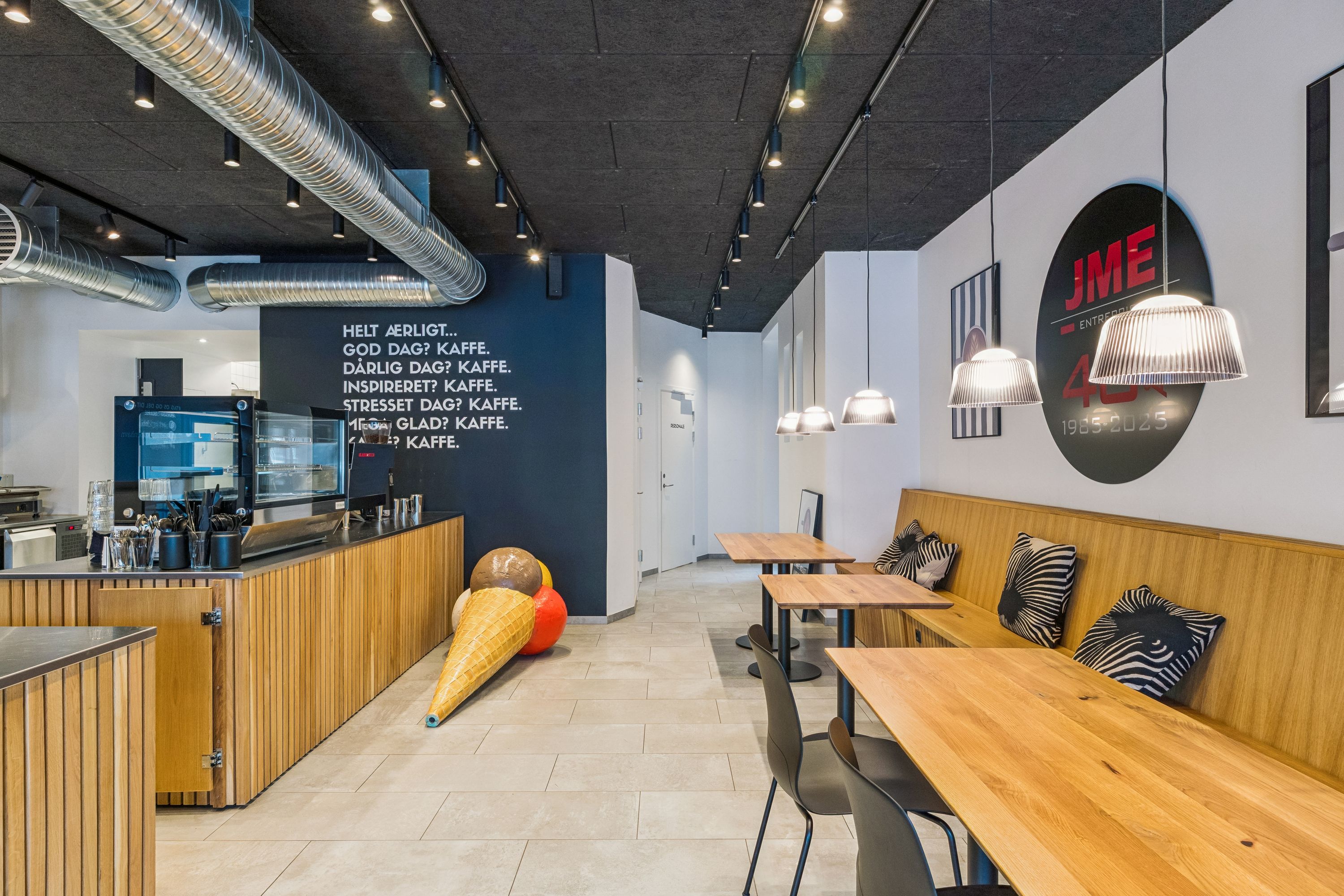 Moderne café med lyse loftslamper, træborde, sofaer og tekst på væggen med citatet Helt ærligt...god dag? Kaffe. Dårlig dag? Kaffe. Inspiriret? Kaffe. Stresset dag? Kaffe.