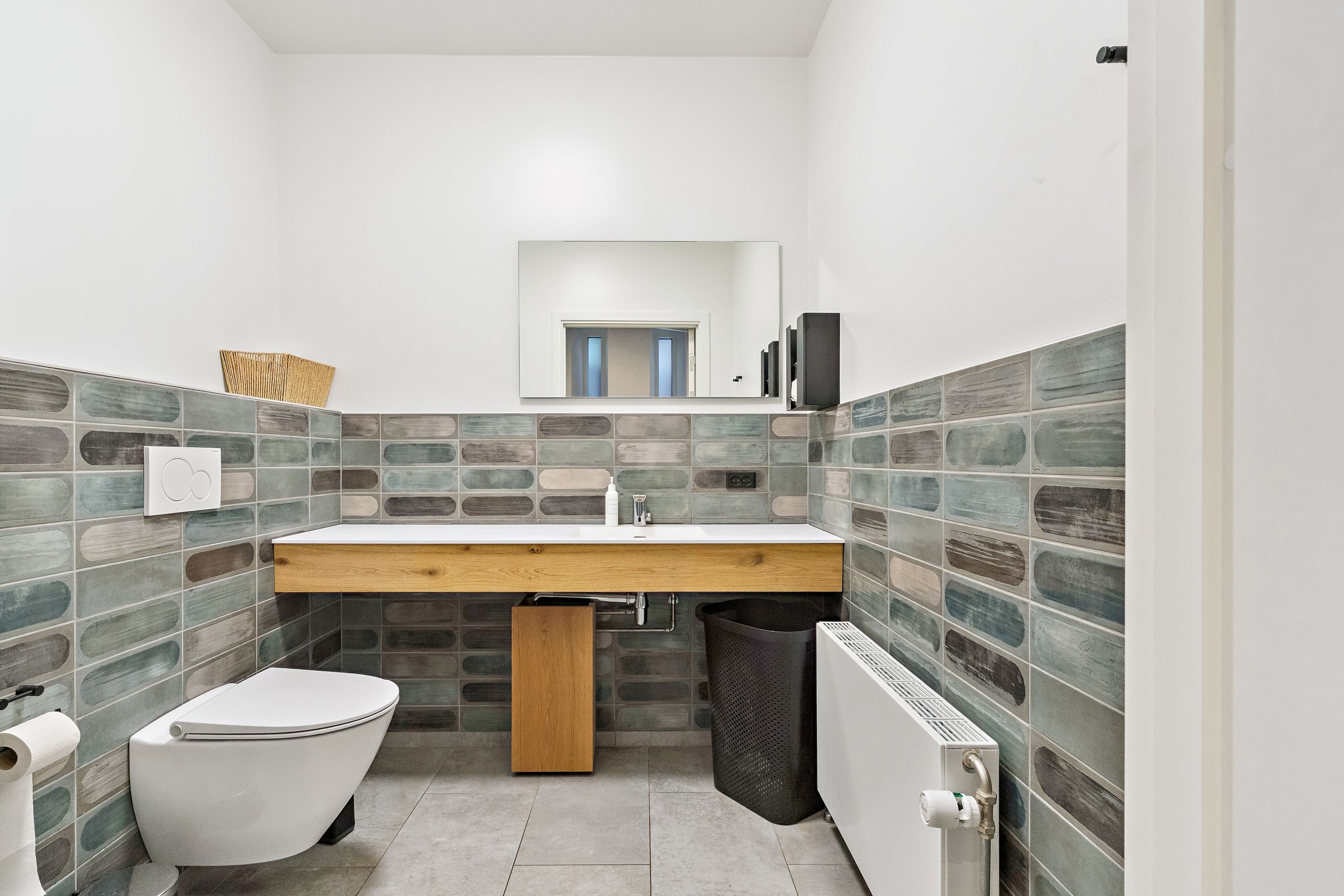 Minimalistisk og moderne badeværelse med lysegrå vægfliser, hvid toilet, tre håndklædetørrere og en stor spejl, der reflekterer rummets lys, samt en hvid radiator og en træplade over vasken