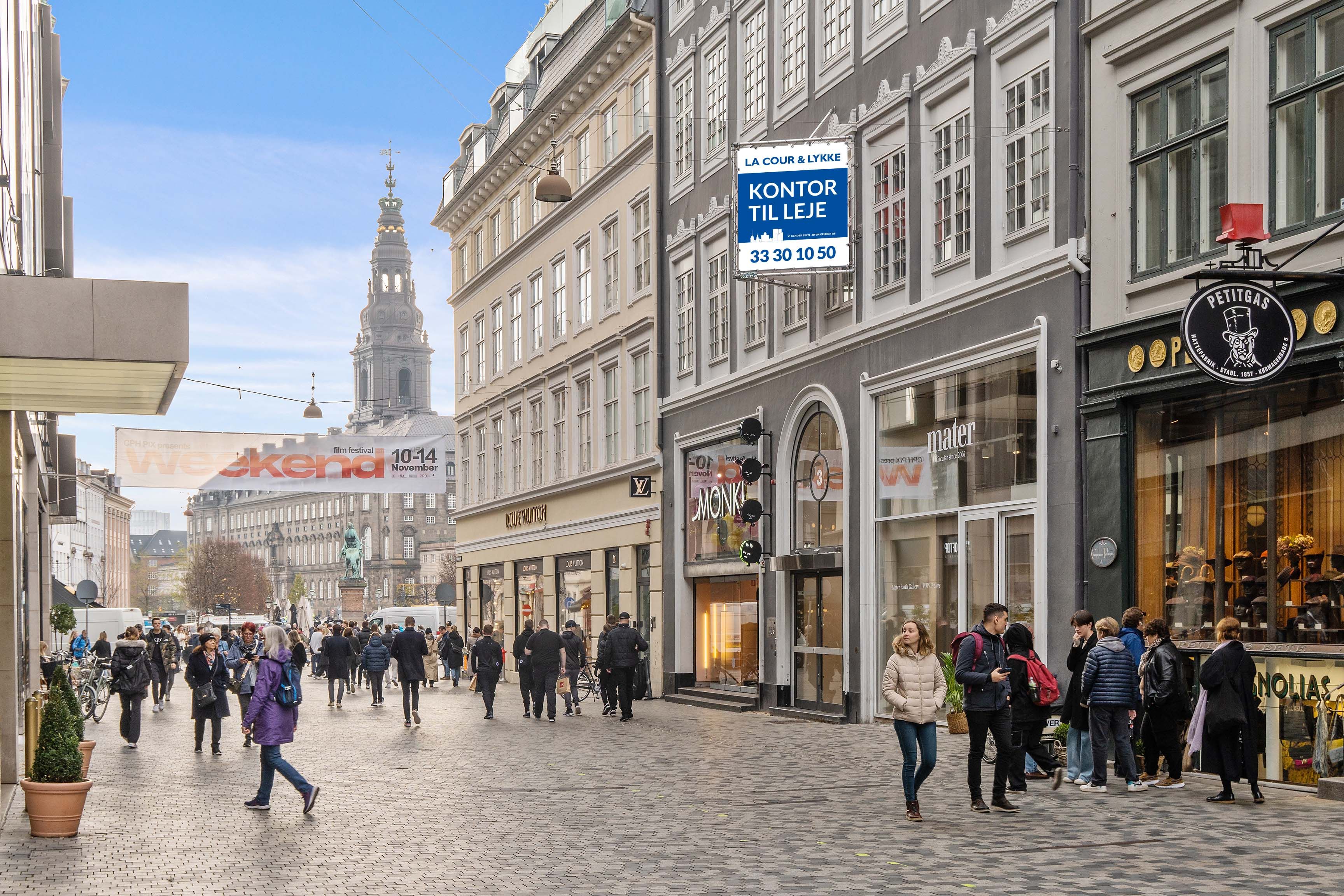 Kontor til leje i København. Folk er på Købmagergade. Kontorskilt med tekst: "Kontor til leje". Der er forretninger med skilte. Bygninger med udskæringer og vinduer.