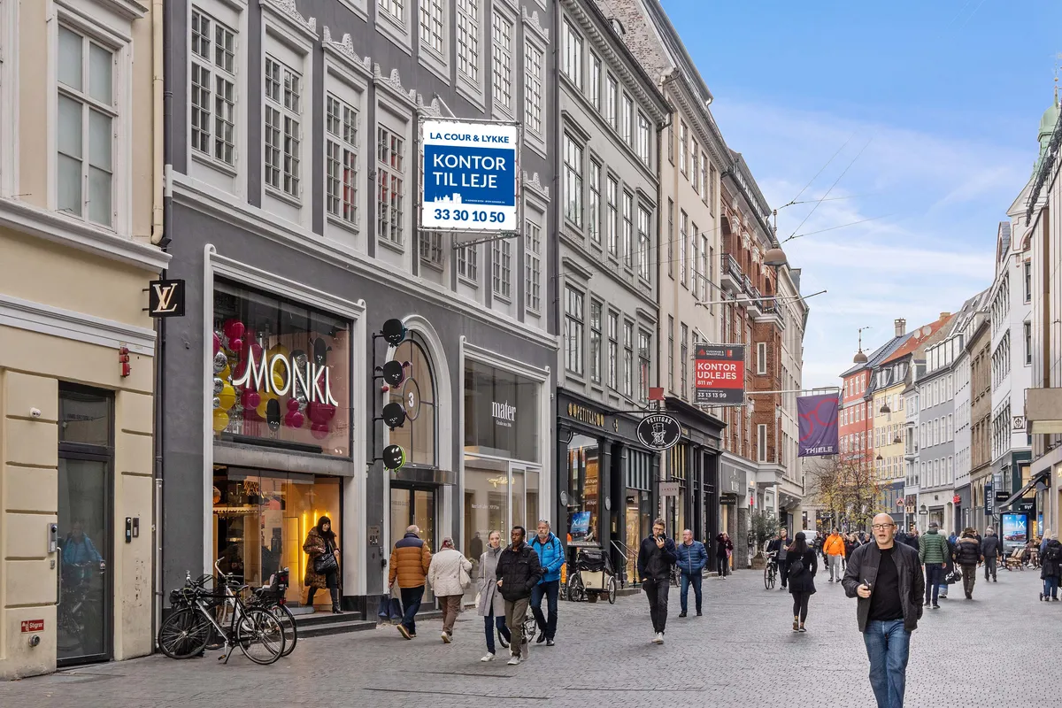 Folk strømmer gennem Købmagergade i København, forbi luksusbutikker som Louis Vuitton og Monki. En stor kontorskilt for La Cour & Lykke er synlig, og det er en lejlighed på 33,30 kvm til leje