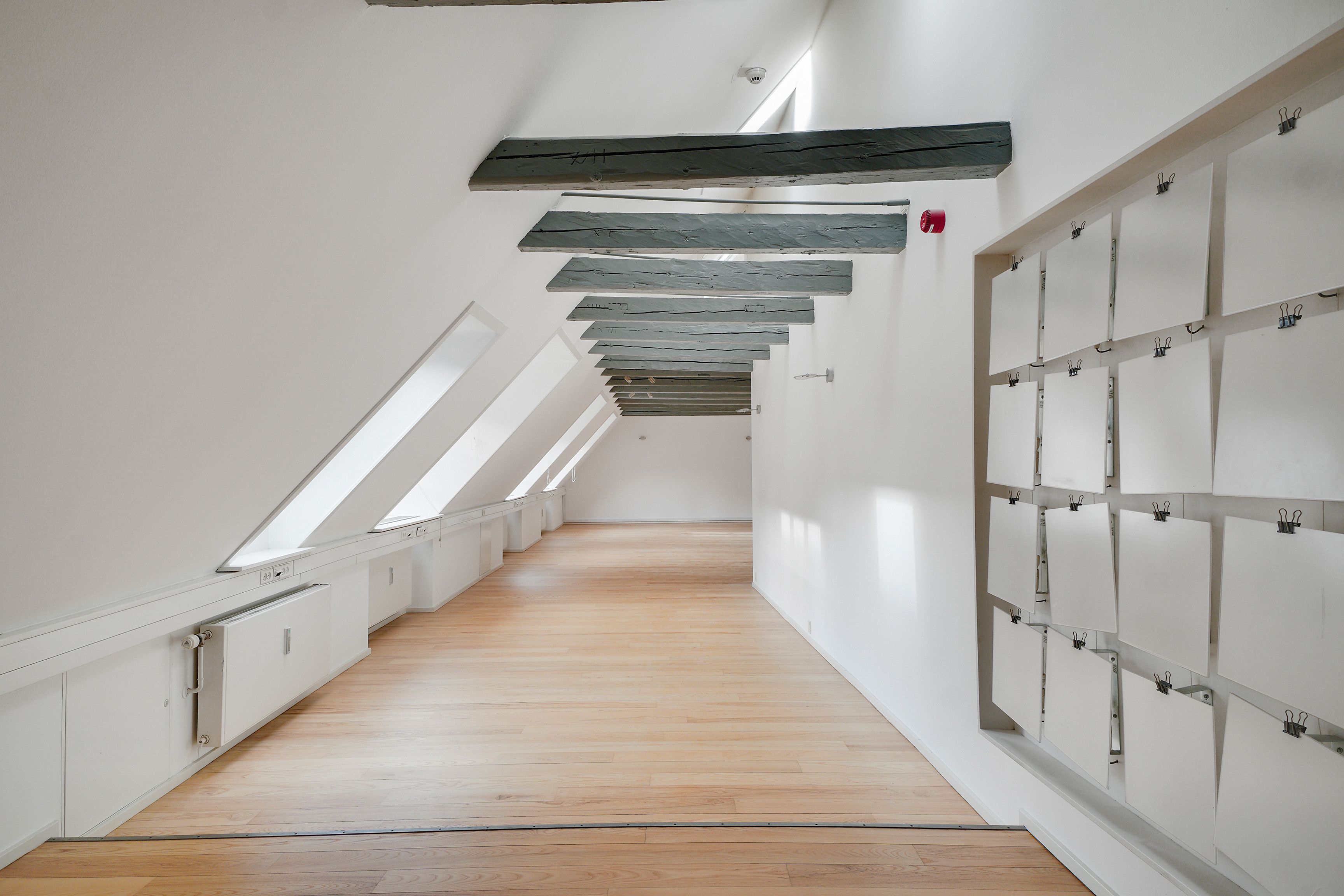 Kontoret har højloft, lyse loftsbjælke og hvid væg. Loftet er dækket af lyseblå træbjælke. Der er hvide vinduer, radiatorer og papirflader med klemmer og huller i væggen.