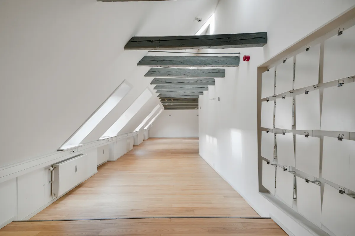 Kontoret har højloft, lyse loftsbjælke og hvid væg. Loftet er dækket af lyseblå træbjælke. Der er hvide vinduer, radiatorer og papirflader med klemmer og huller i væggen.