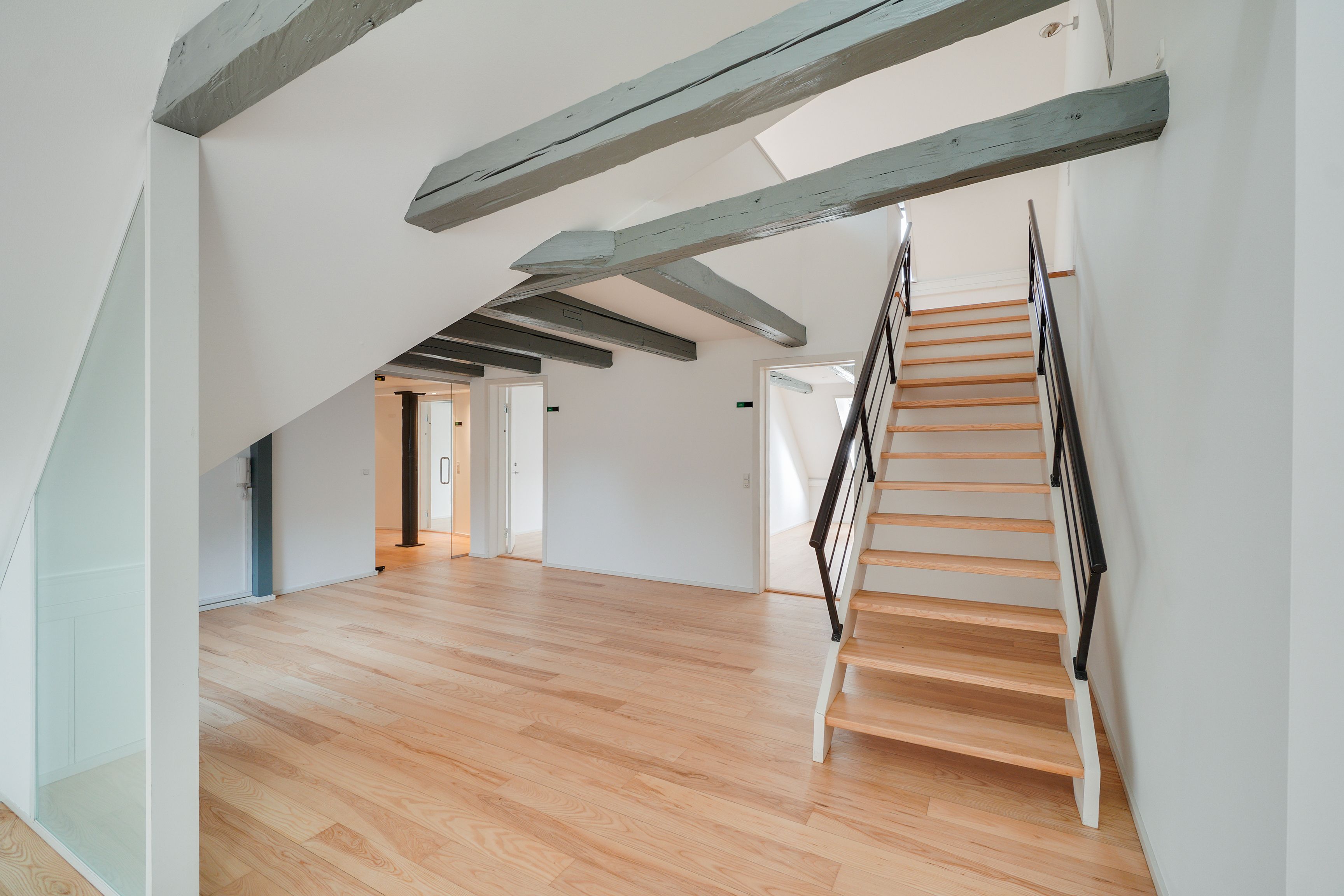 Kontor med åbent loft og trægulv i lyse farver. Stiger med håndtag fører op til næste etage. Lukket dør og glasdør med håndtag i baggrunden.