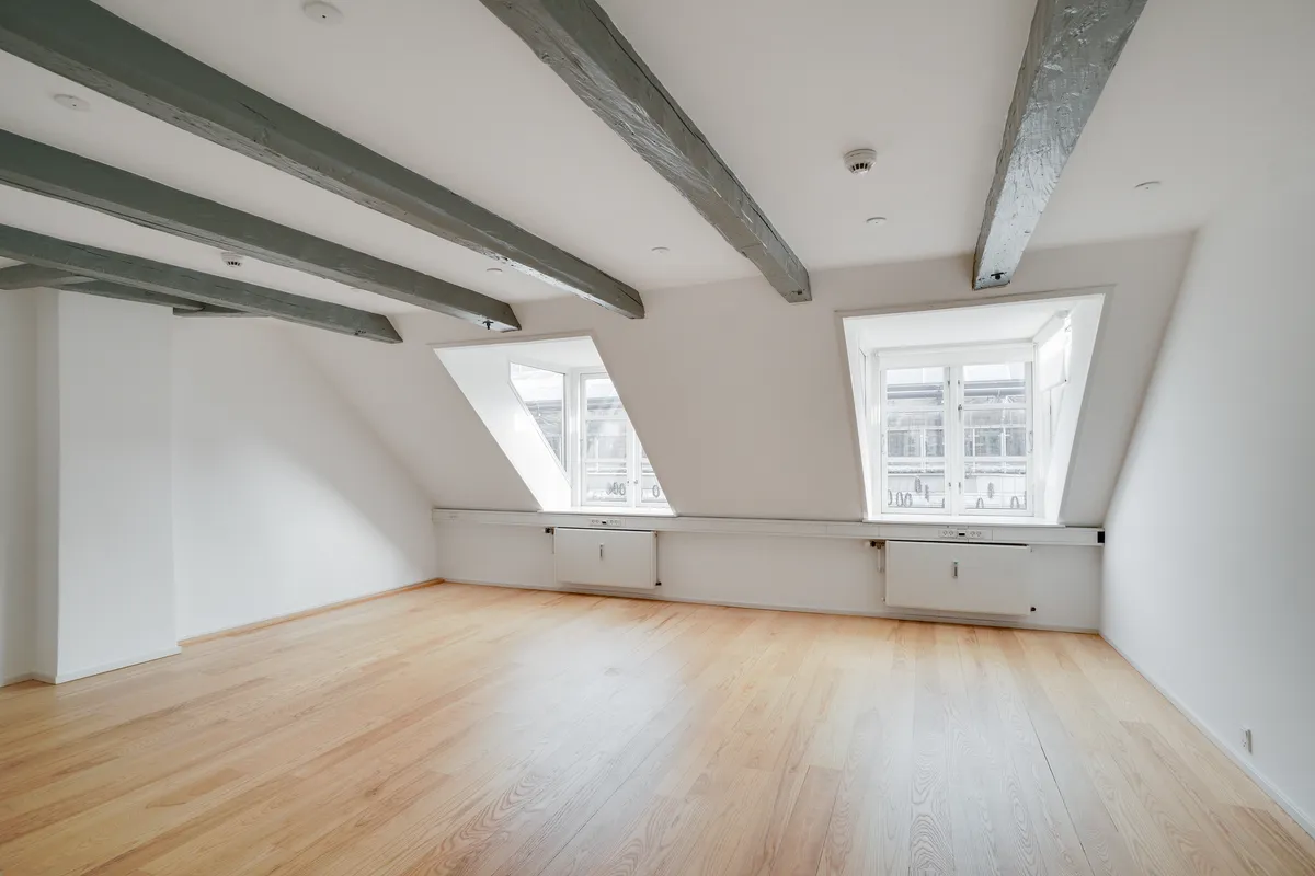 Kontor med højloft, parketgulv, hvide vægge og loftsvinduer. Højloft med træbjælker og hvide vægge. To store vinduer med hvide rammer. Ingen møbler eller genstande i rummet.