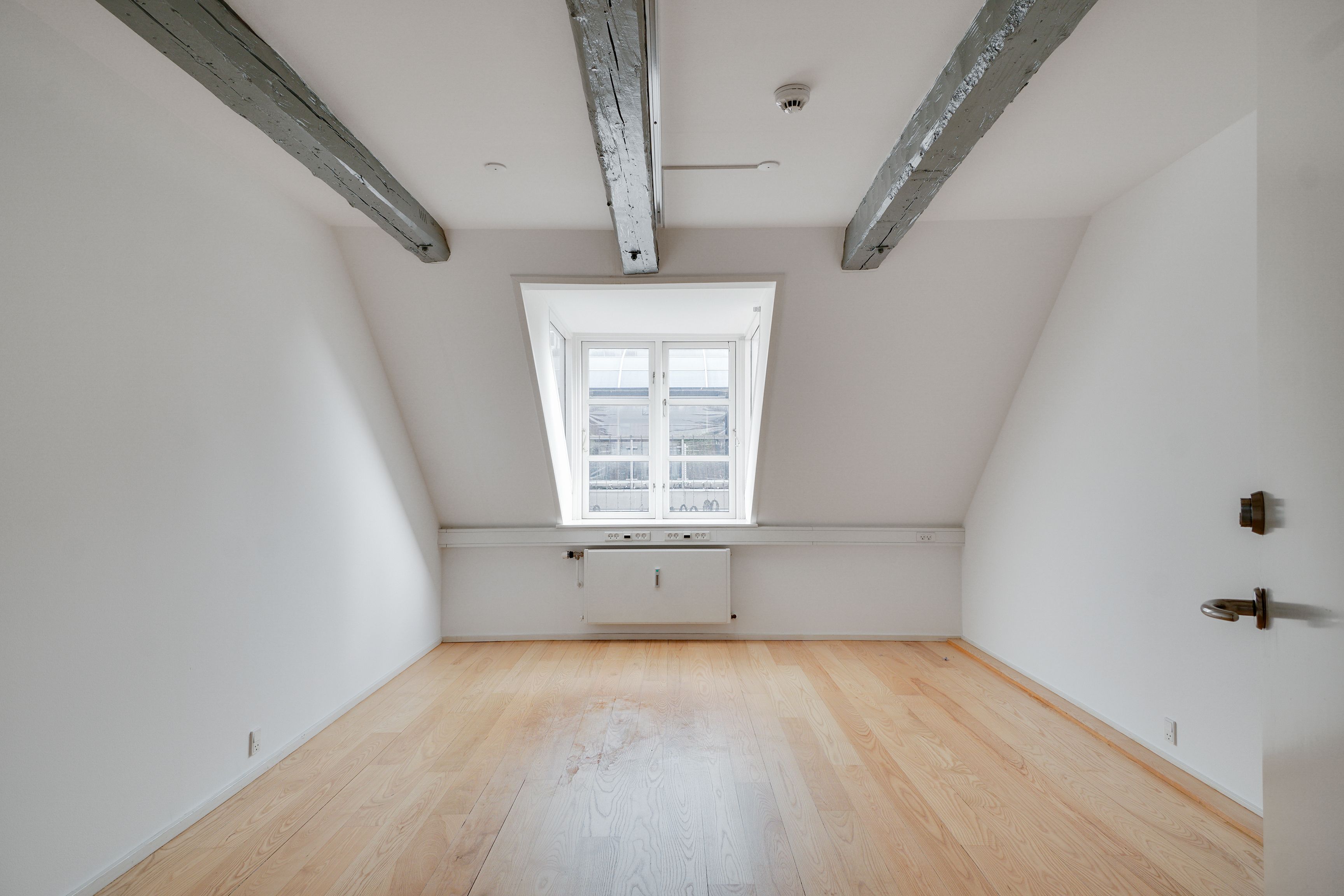 Lej kontor i lyse loftsrum med hvide vægge og trægulv i København. Rummet har lyse træbjælker, lys og vinduer og et hvidt radiatorsystem