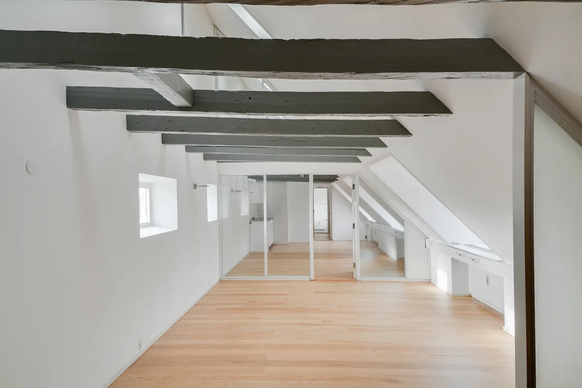 Minimalistisk design med hvide vægge, lyse trægulve og slanke bjælker, der giver loftet et varmt og indbydende look, samt en smal gang med spejle og vinduer