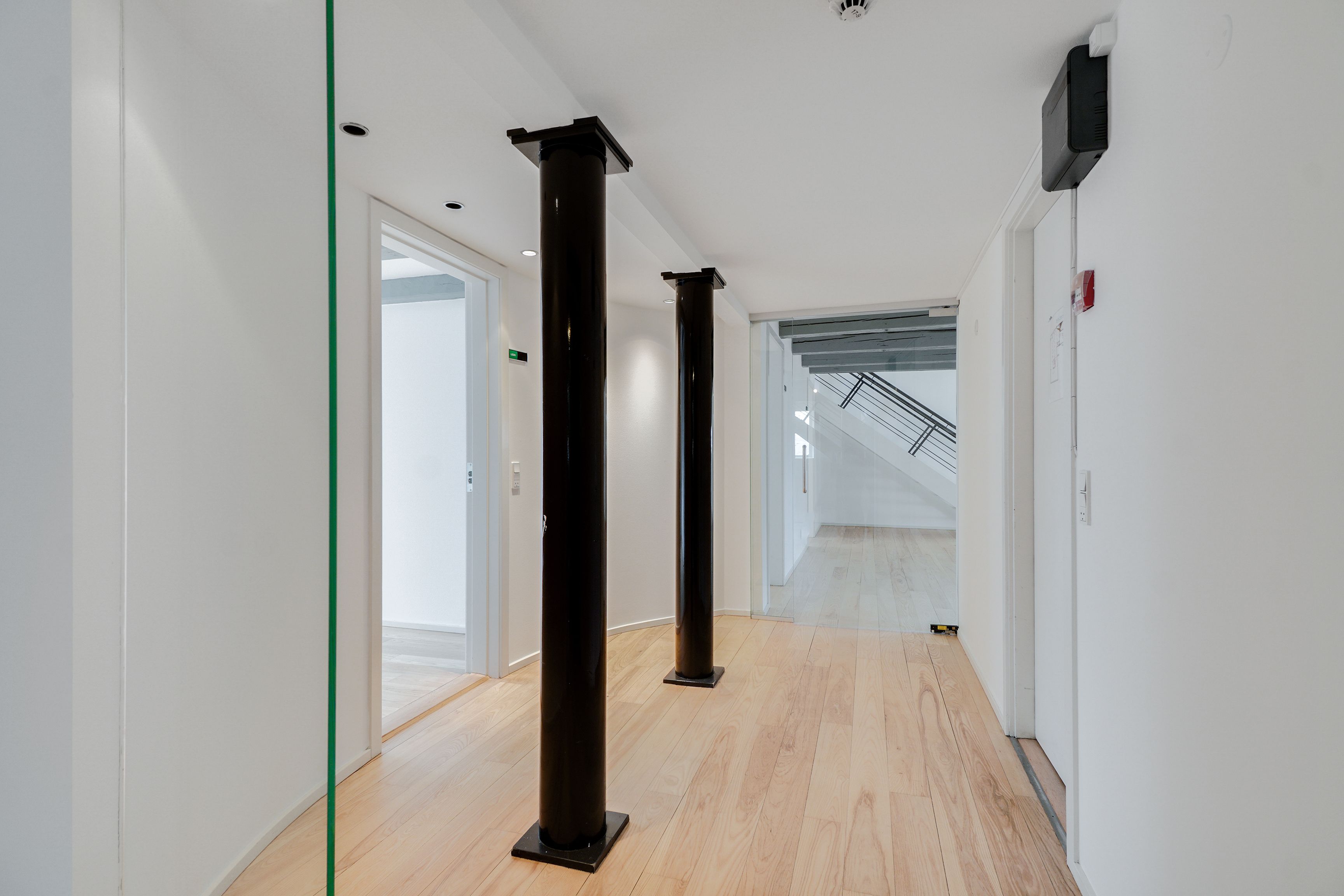 Lækkert kontor med højloft og lyse gulve i hjertet af København. To store sorte søjler og moderne møbler. En åben dør og et vindue leder ind i en åben stue.
