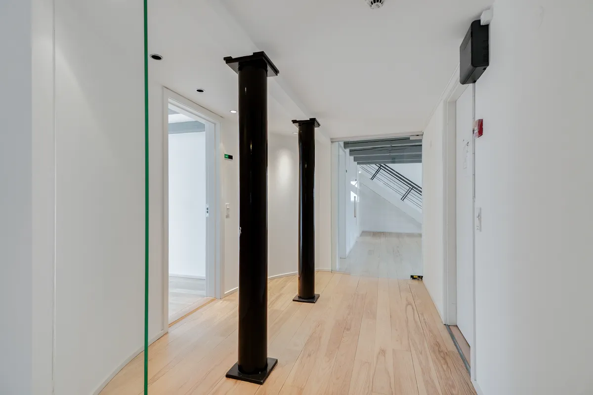 Lækkert kontor med højloft og lyse gulve i hjertet af København. To store sorte søjler og moderne møbler. En åben dør og et vindue leder ind i en åben stue.