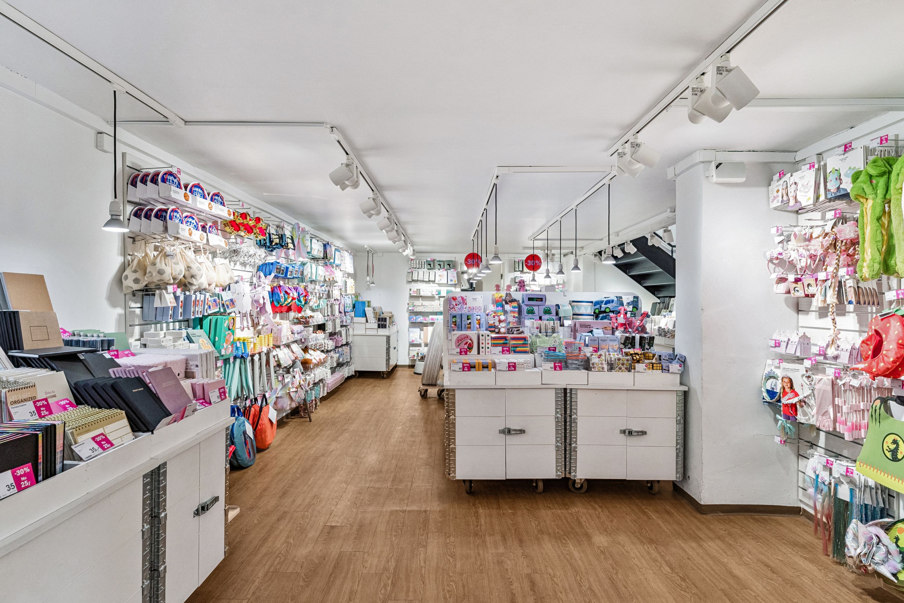 En moderne hvid indretning med lyse gulve, flere hylder med legetøj og papirvarer samt flere rabatter og produkter i salg.