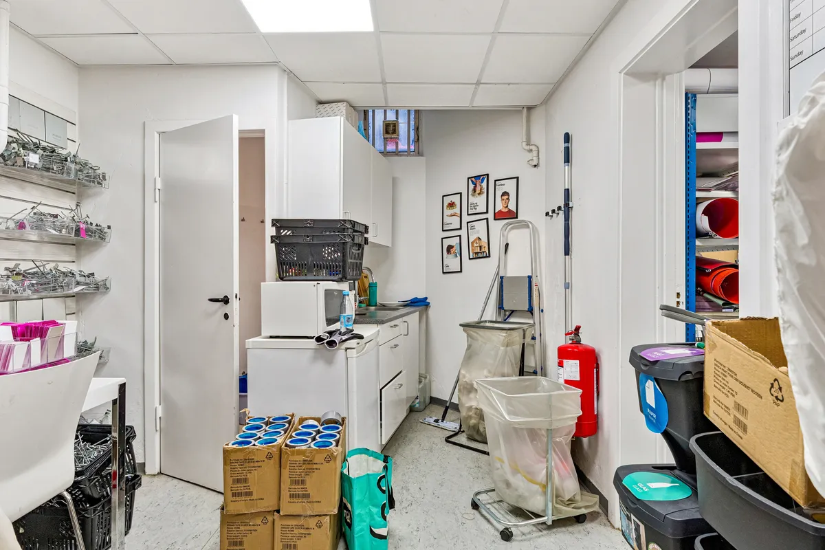 Køkken med hvide vægge, gulv og møbler, tomme opvaskmaskine og mikroovn, tøjstativ, rengøringsprodukter, papkasser og plastikposer på gulvet og væggen, og flammeværn på væggen