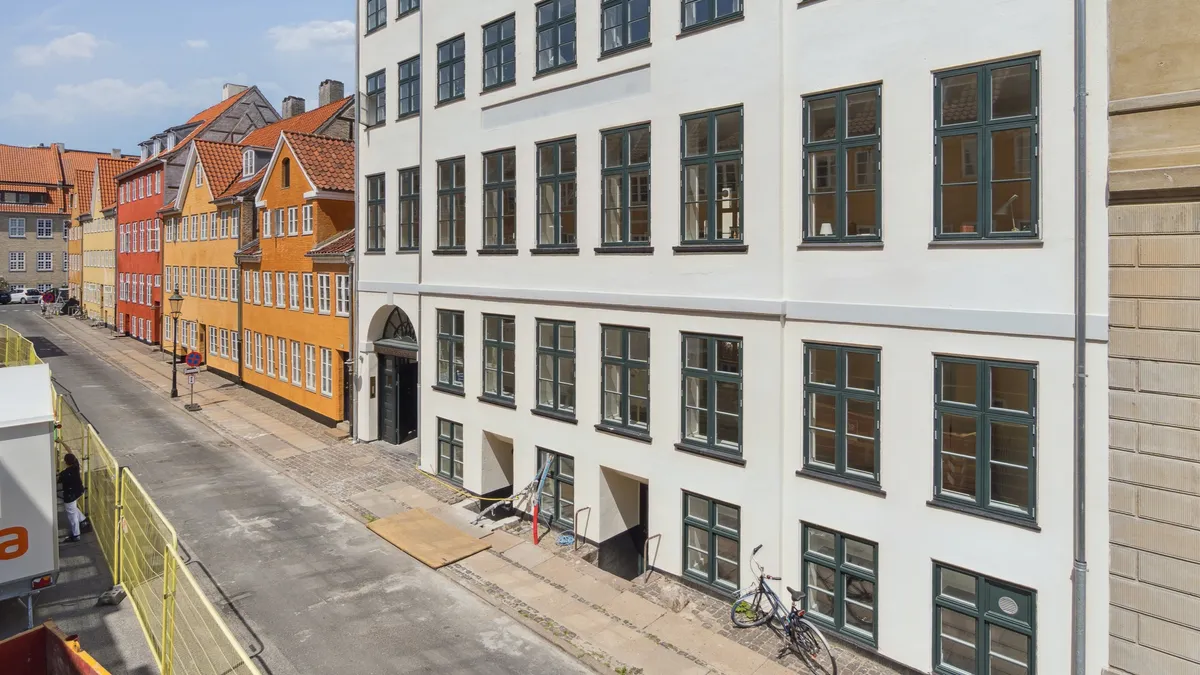 Hjørnebygning med højde, vinduer og cykel på fortovet. Bagved er byggekran og personer i arbejde. Huse i røde og gule farver.