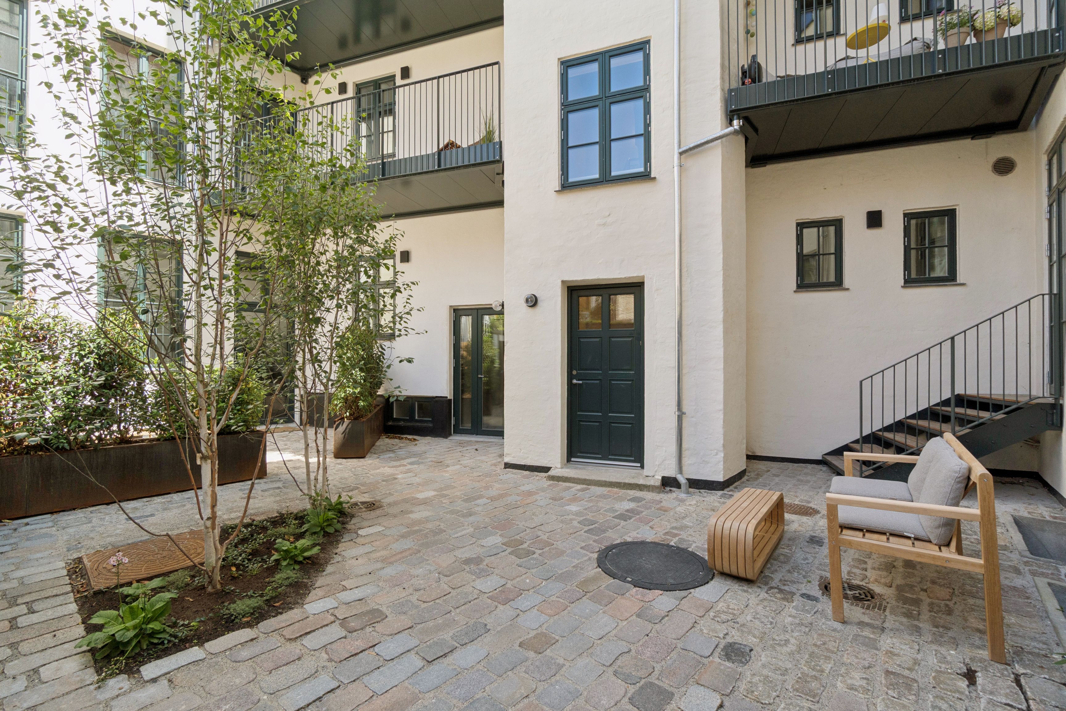 Moderne hjørne lejlighed med have adgang, terrasse og lyse gulve. Møbler og kunstværker på terrassen. Lejlighedsdør og balkon med plantepotte.