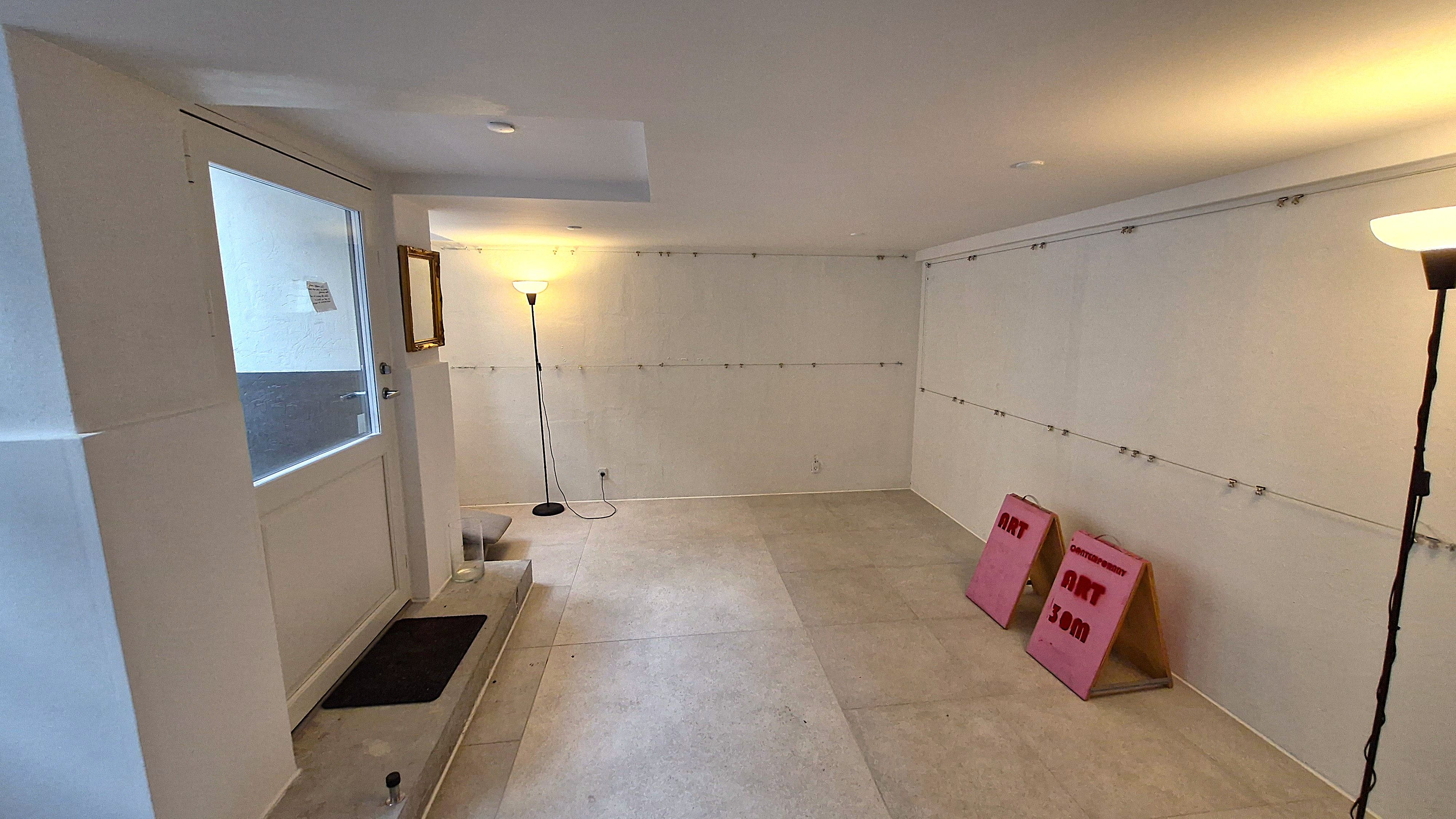 Et rum med hvide vægge og gulv, to pink skilte, en lysestage og en spejl i kortet, en dør med en sort gulvtæppe og en åben dør