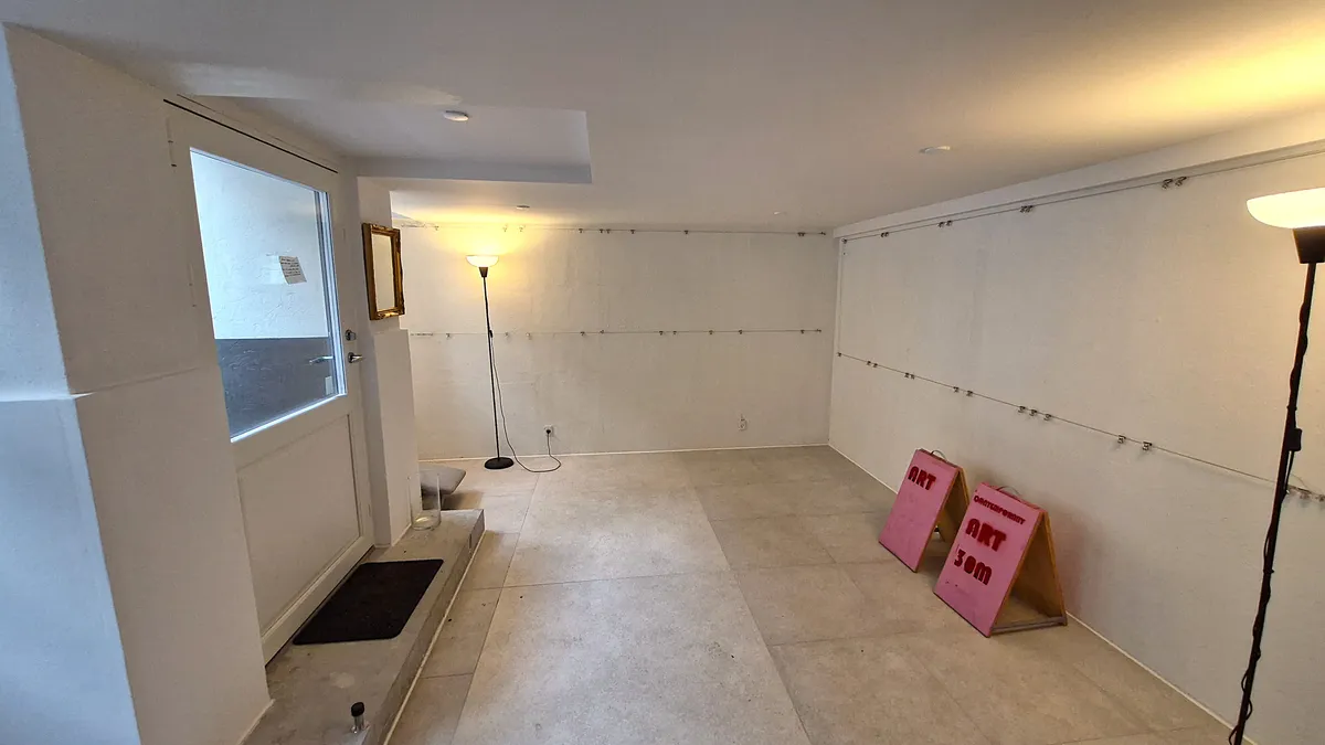 Et rum med hvide vægge og gulv, to pink skilte, en lysestage og en spejl i kortet, en dør med en sort gulvtæppe og en åben dør