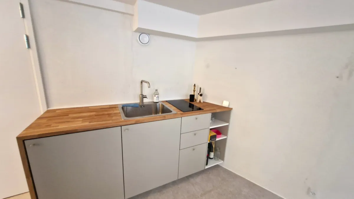 Et lille køkken med rustfrit stål skab og vask, hvid væg og træbenk, samt induktionskomfur og håndvaskekran.