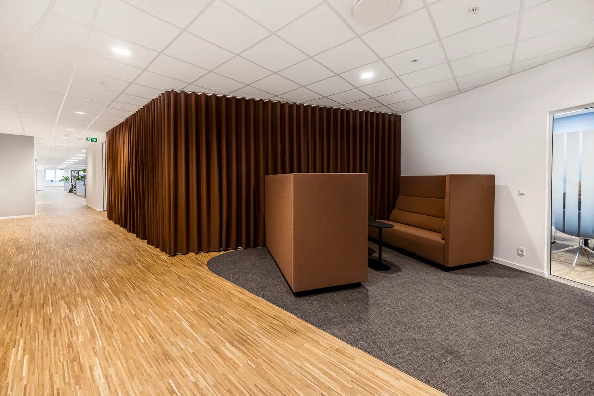 Kontorrum med moderne design og fællesfaciliteter. Brune lydabsorberende vægge og trægulv. Sovesofa og mødebord i lysbrunt læder. Kaffebord og lysegule loftslys.