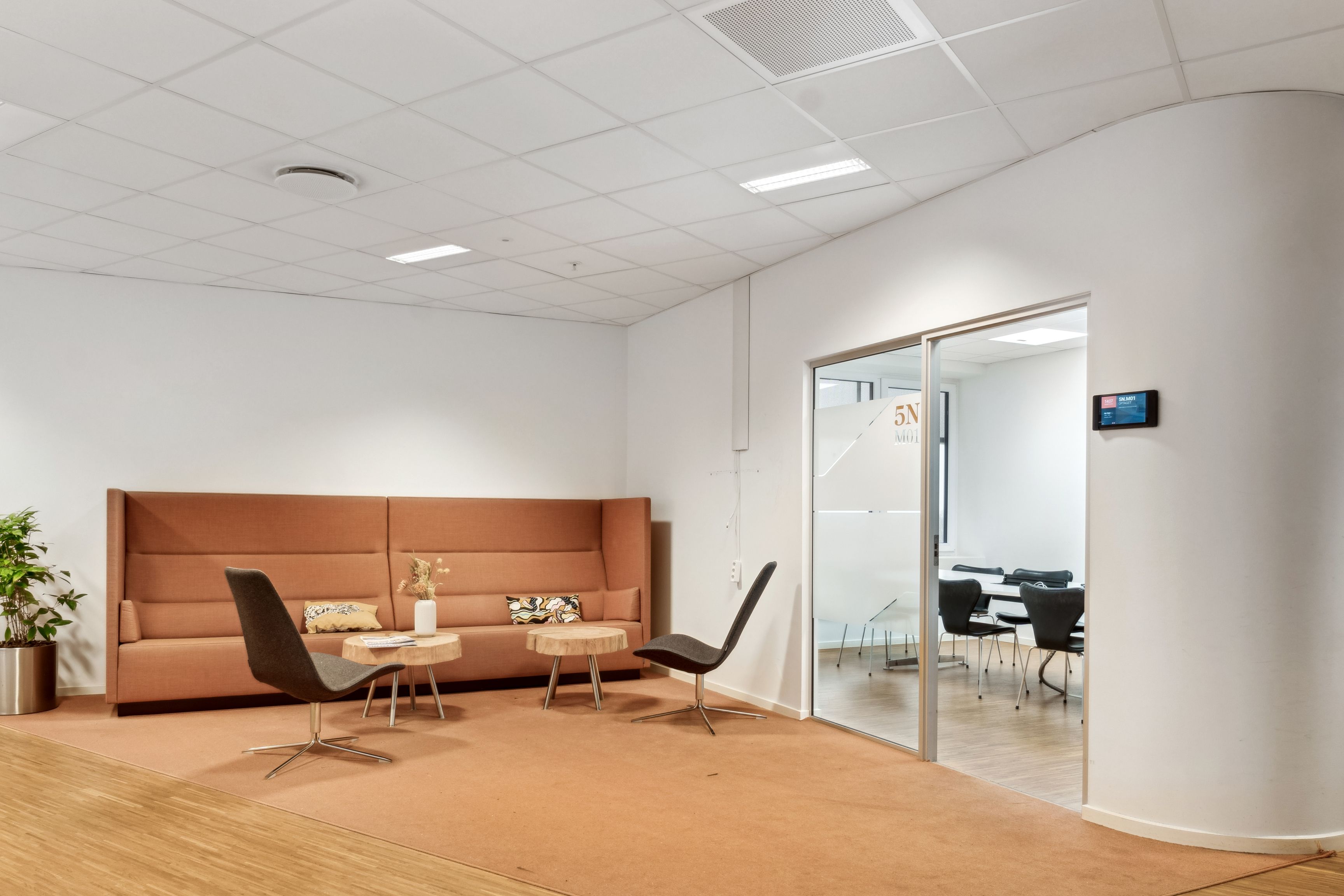 En moderne kontorlounge med lyse hjørner og en beige sofa. To stole med lyse beige sæder og høje ben står på hver side af sofaen. Et skrivebord med en plante og en lampe.