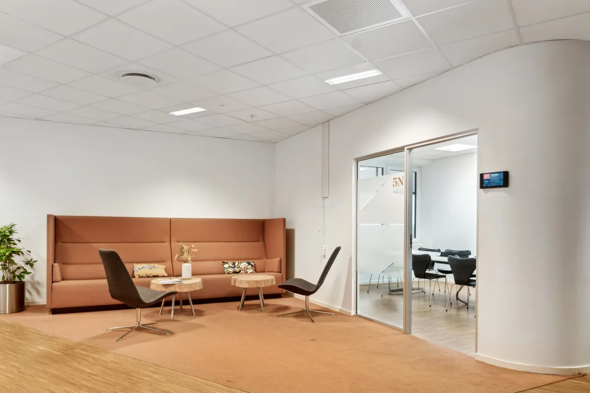 En moderne kontorlounge med lyse hjørner og en beige sofa. To stole med lyse beige sæder og høje ben står på hver side af sofaen. Et skrivebord med en plante og en lampe.