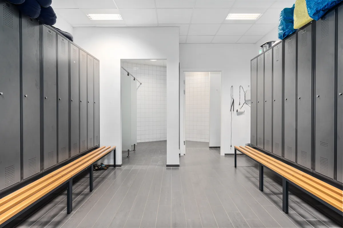 En stor, lyse og moderne garderobehal med to lange bænke og flere garderobeskabe med tøj og andre genstande. Bagved er der flere døre og et lukket badeværelse.