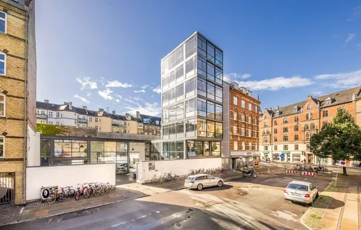 En moderne bygning med store glasruder og flere parkerede biler og cykler i en bydel med gamle huse og træer